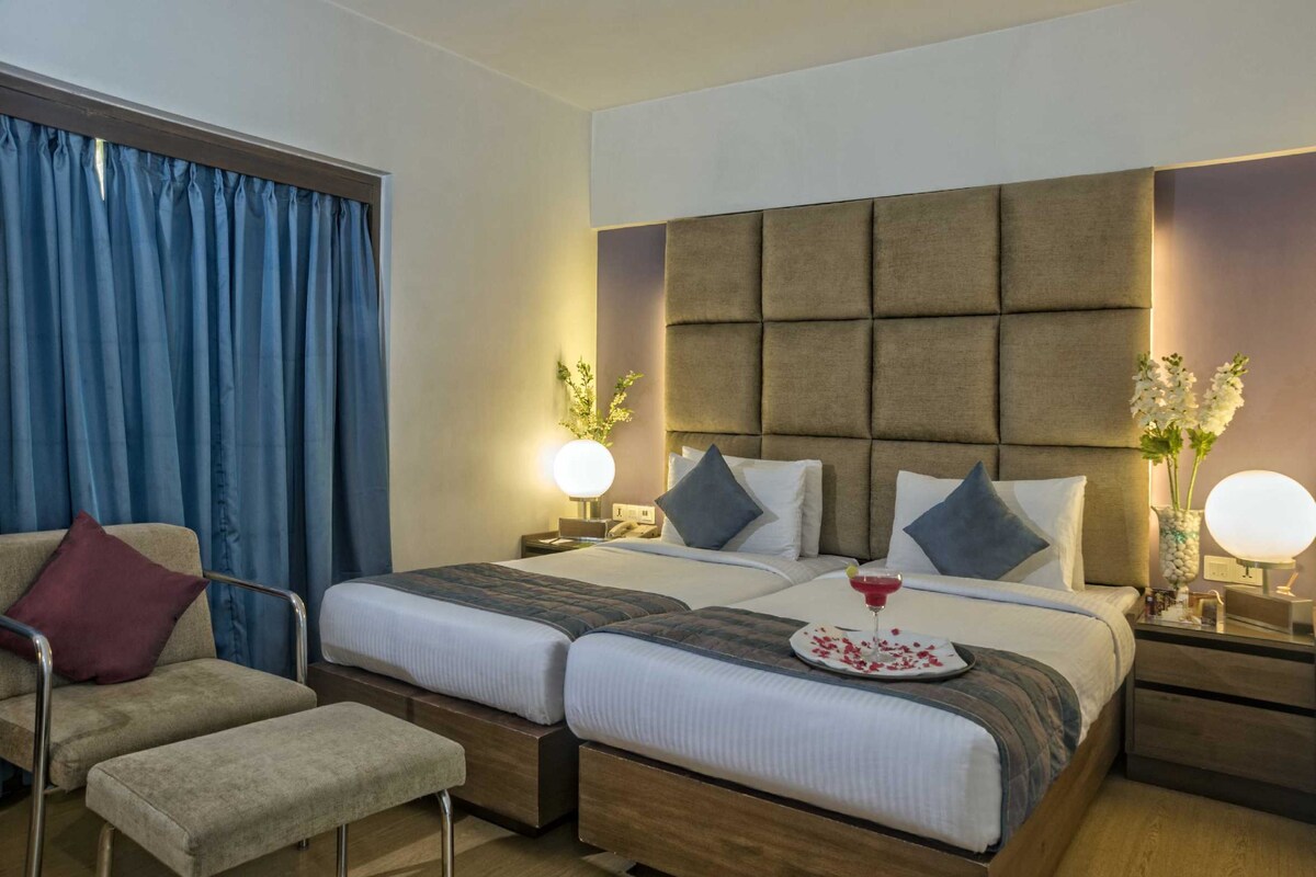 A hotel room with two beds, a sofa, and a chair. There are bedside tables with lamps and a decorative vase.