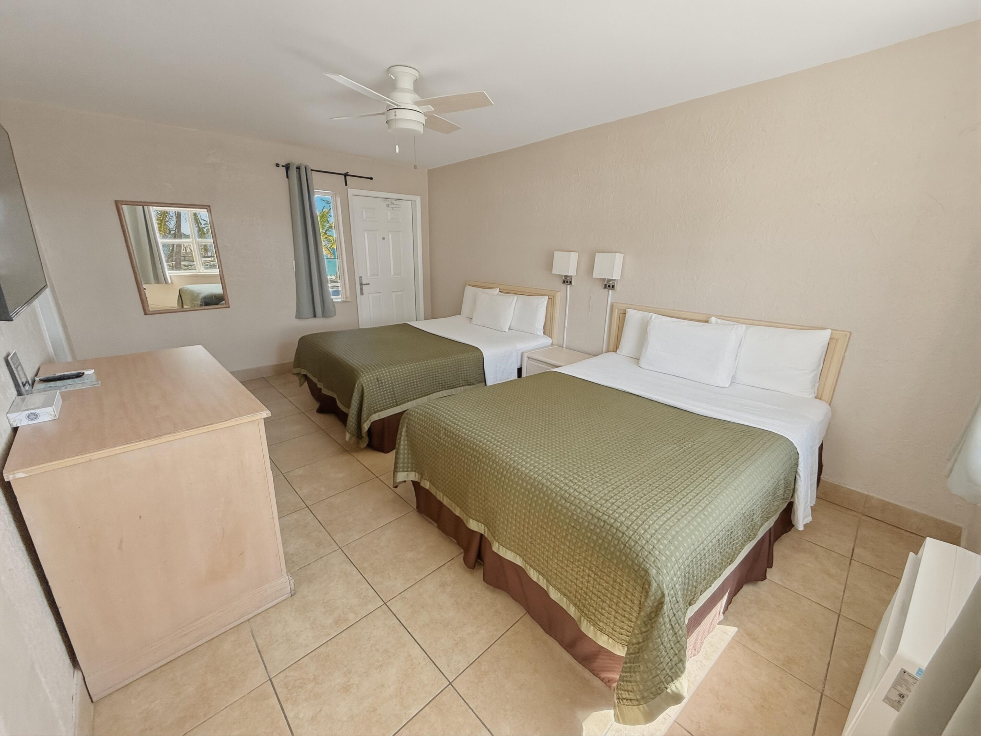 A hotel room with two beds, a desk, a ceiling fan, a TV, and a window with curtains.