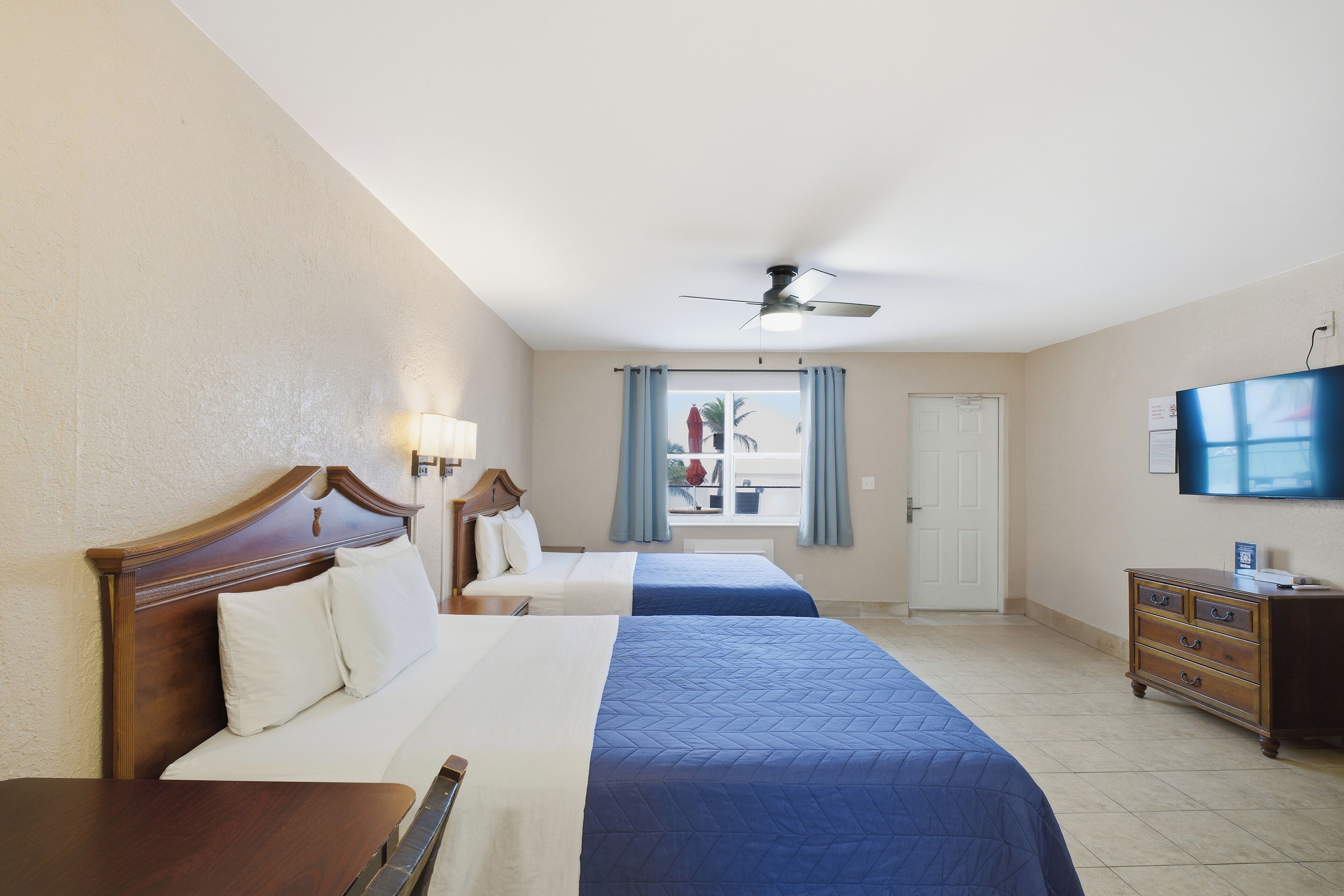A hotel room with two beds, a ceiling fan, a television, and a window with curtains.