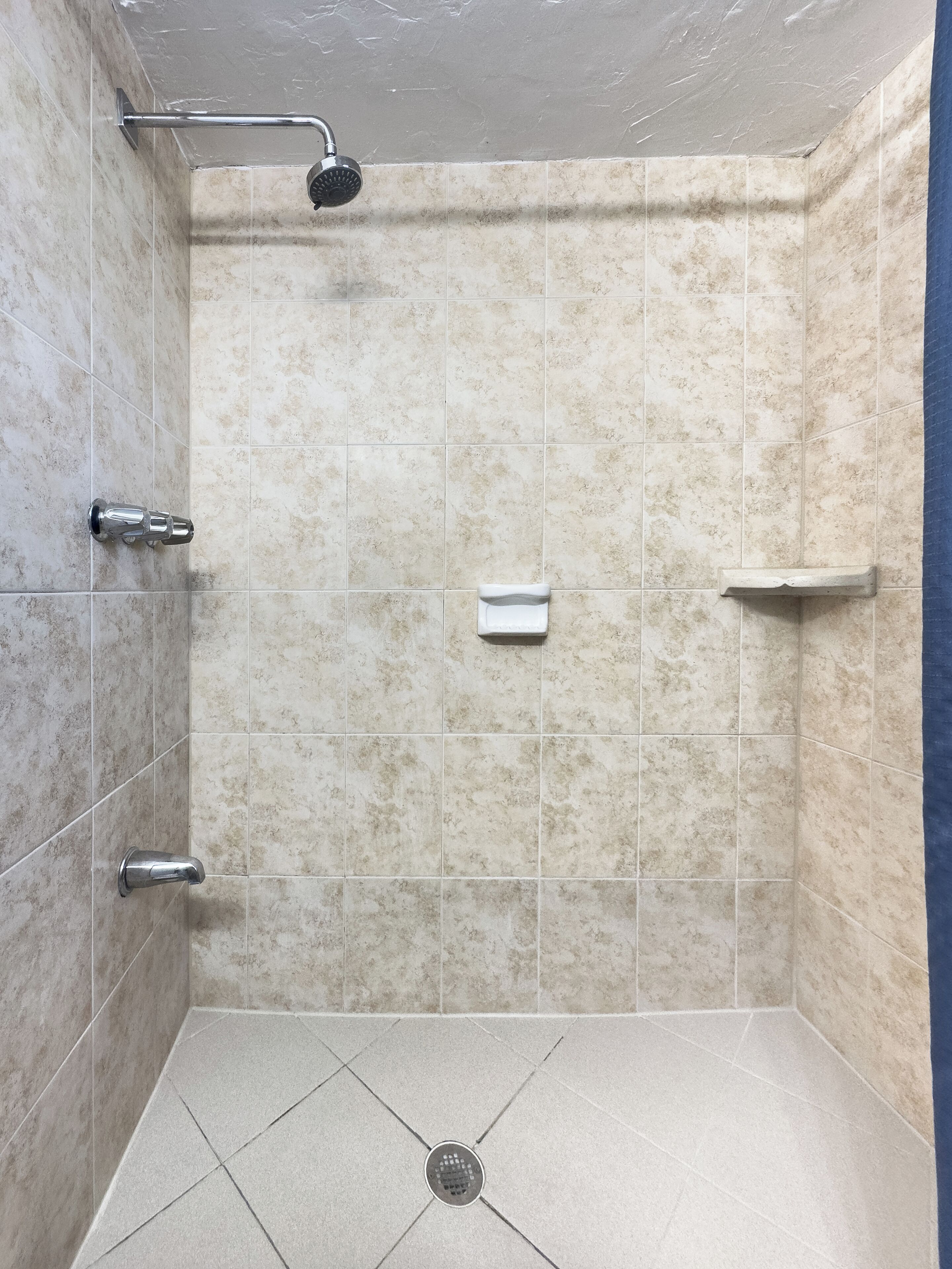 A small, tiled shower room with a handheld showerhead, a handheld faucet, and a built-in shelf.