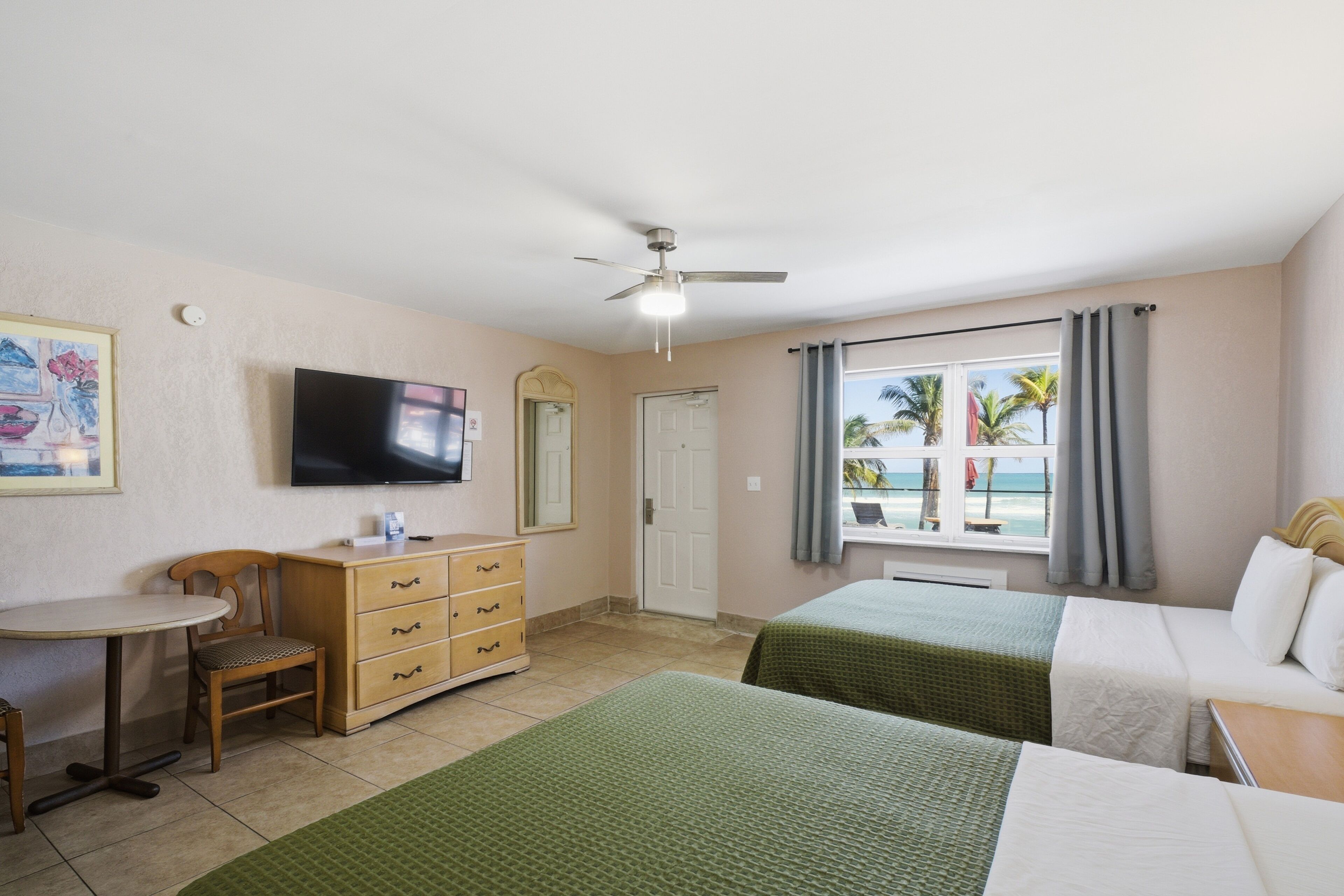 A hotel room with two beds, a TV, a desk, and a window with a view of palm trees and the beach.