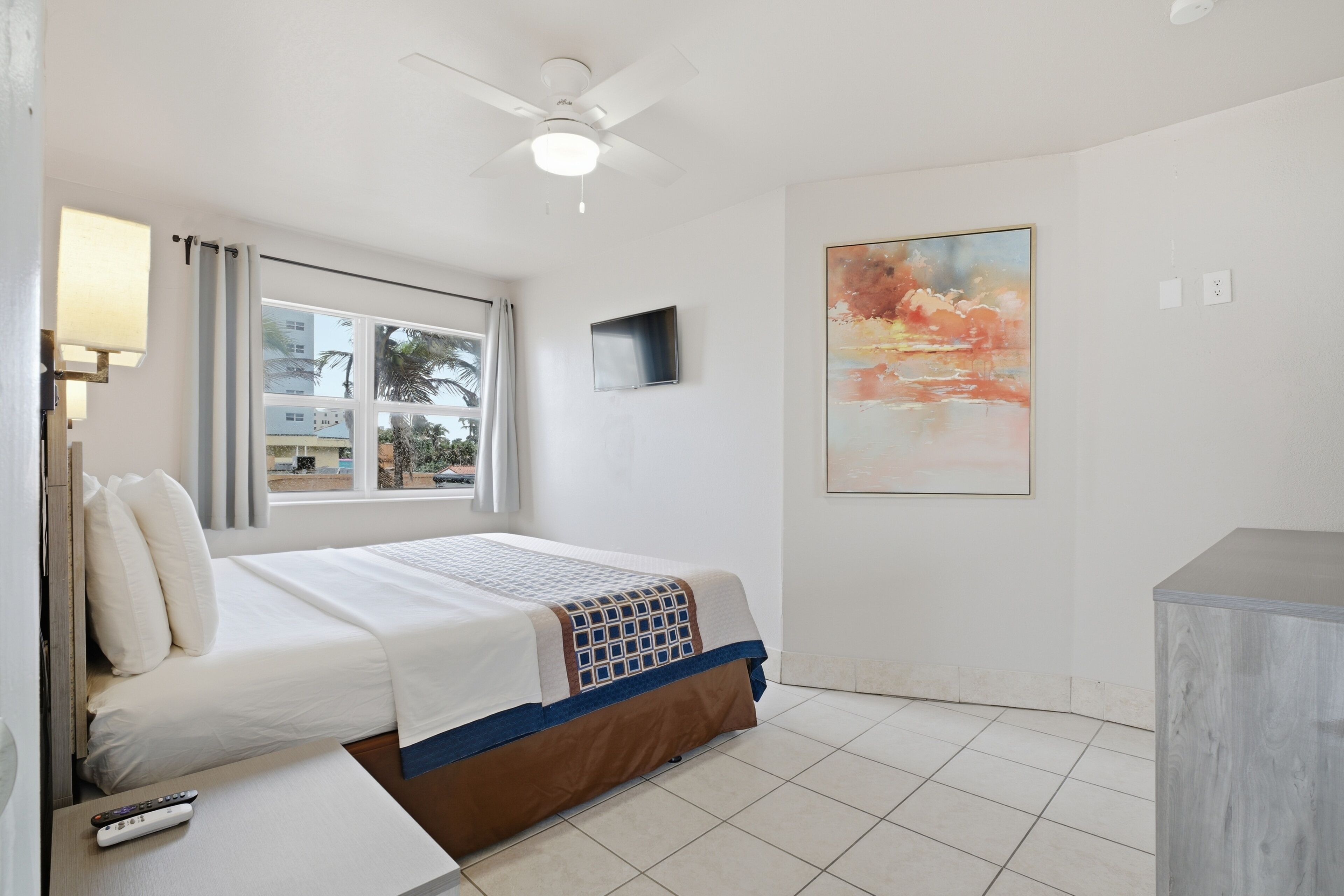 Una habitación de hotel con cama, ventilador de techo, televisión, un cuadro y ventana con cortinas.