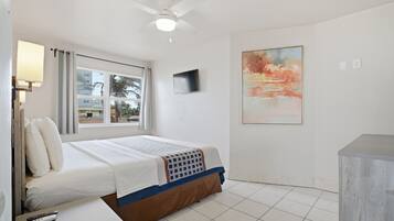 Una habitación de hotel con cama, ventilador de techo, televisión, un cuadro y ventana con cortinas.