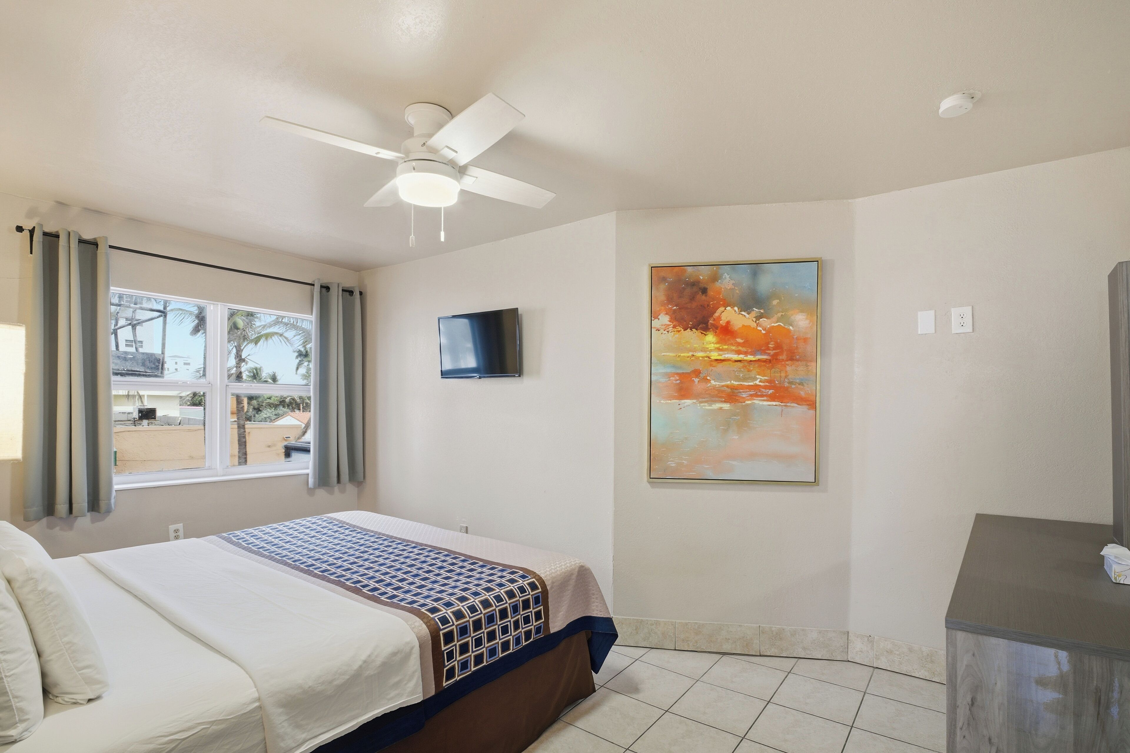 A hotel room with a bed, a ceiling fan, a TV, a painting, and a view of the outside.