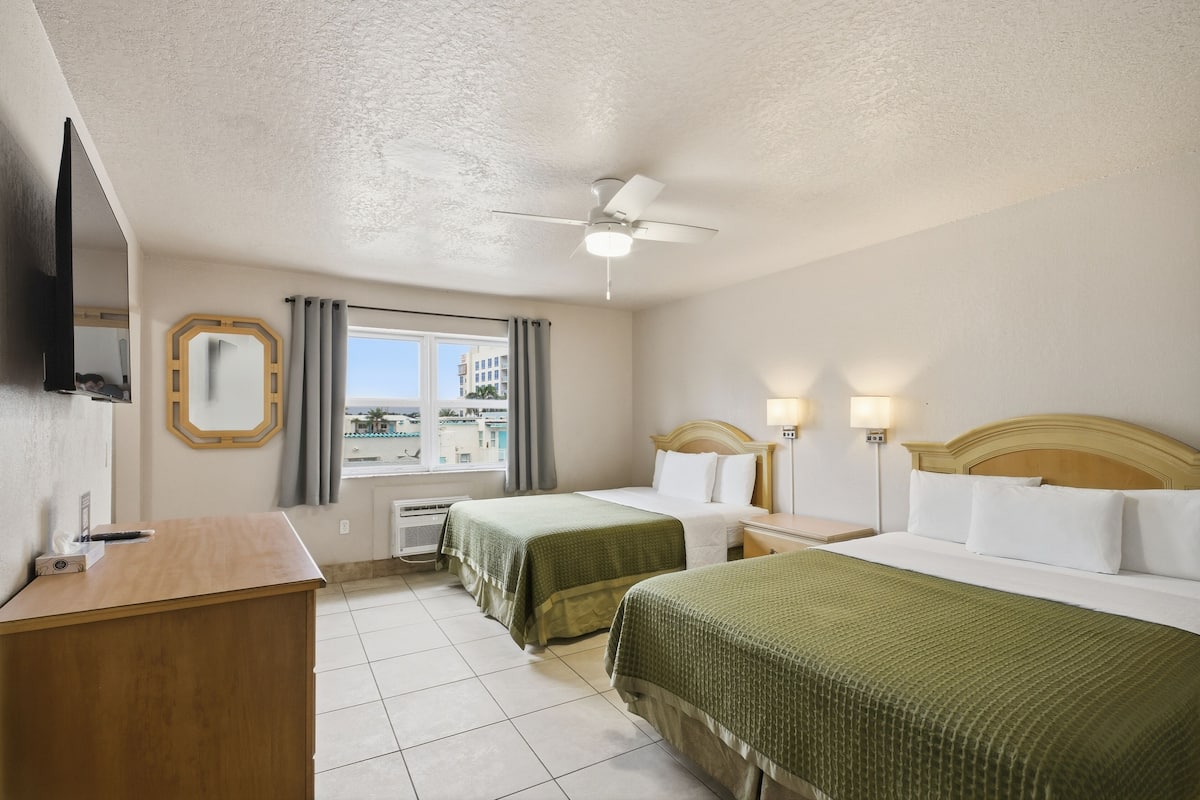 A hotel room with two beds, a desk, a television, a window with curtains, and a ceiling fan.