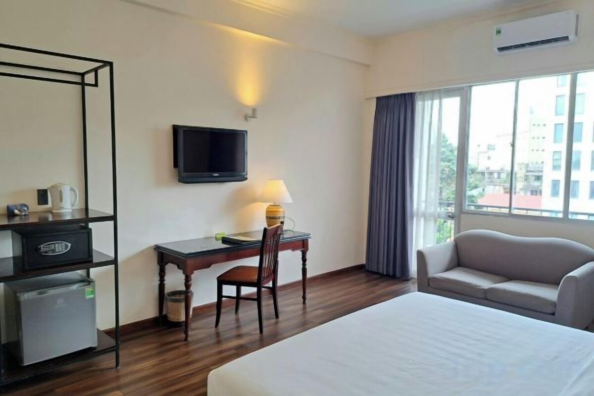 A hotel room with a bed, a desk with a chair, a television, a sofa, and a window with curtains.