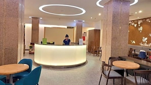 Un vestíbulo de hotel con una mesa de recepción, sillas azules, mesas de madera y una pared decorativa con motivos florales.