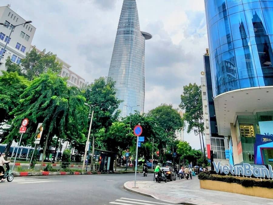 Uma rua urbana moderna com um edifício de vidro imponente, uma faixa de pedestres e áreas verdes ao redor.