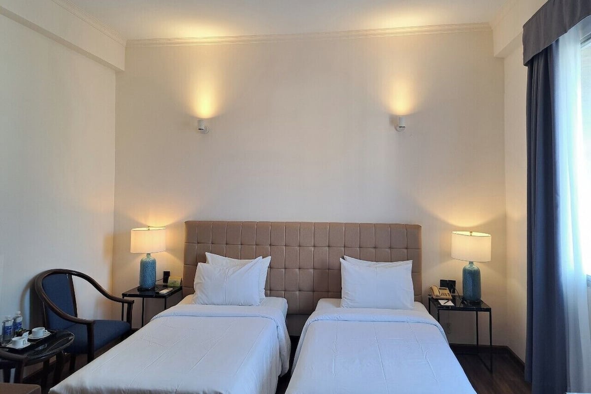 Hotellihuone, jossa on kaksi sänkyä, sängynpääty, yöpöytävalaisimet, tuoli, pieni pöytä ja ikkuna, jossa on verhot.