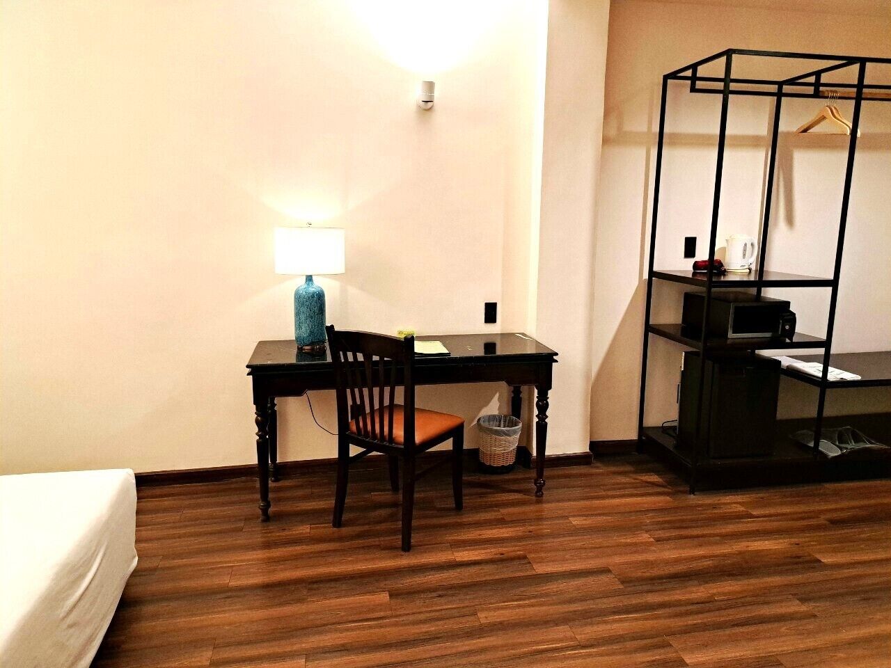 A room with a wooden floor, a desk with a chair, a lamp, a hanging shelf, and a bed.