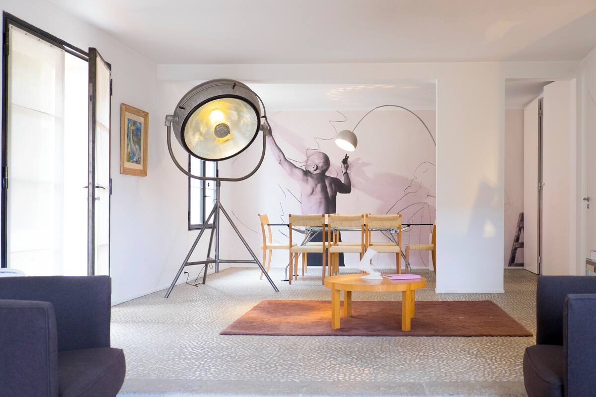 Een moderne woonkamer met een grote vloerlamp, een houten tafel en een stoel. Er hangt een schilderij aan de muur en er staat een ladder in de hoek.