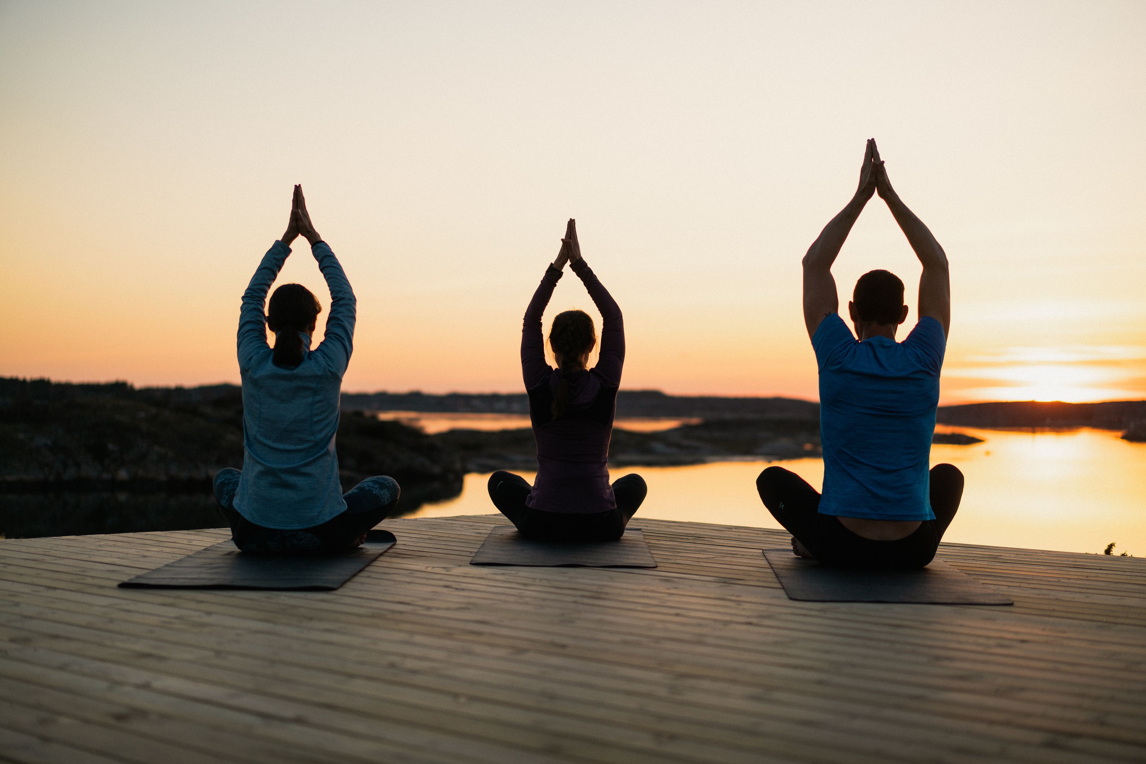 Three people performing yoga on mats on a wooden deck with a sunset view.