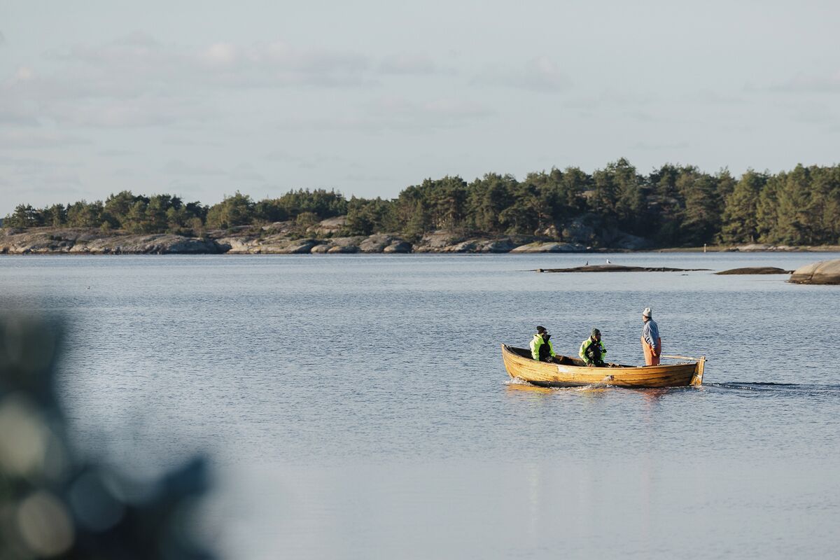 A wooden boat with three people on a calm body of water, surrounded by a forested shoreline.