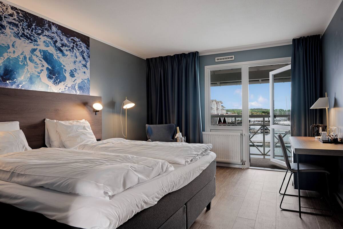 A hotel room with a large bed, a desk with a chair, a view of a balcony, and a painting on the wall.