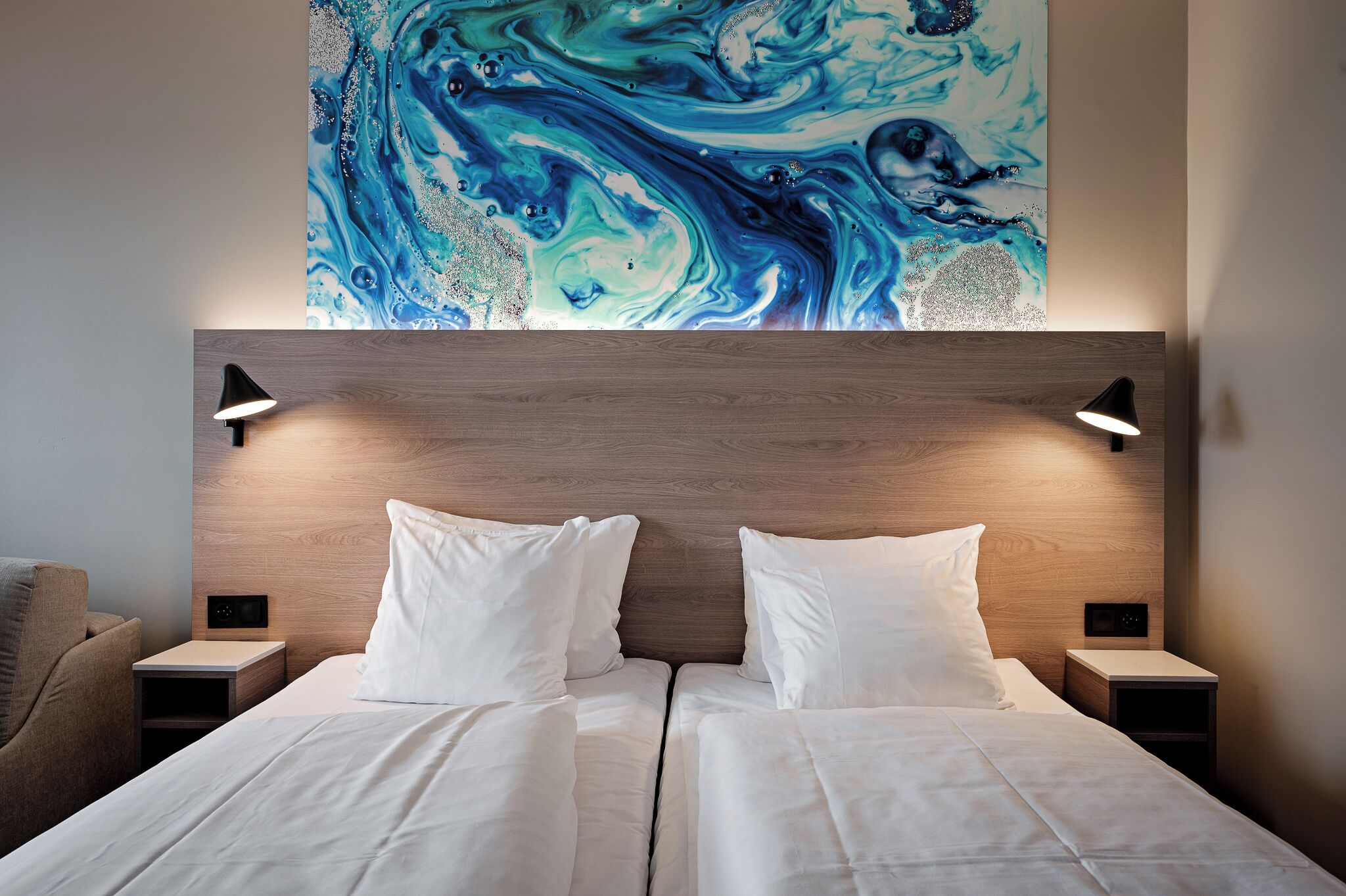 Una habitación de hotel con una cama grande, dos mesitas de noche con lámparas, un cabecero y una obra de arte en la pared.