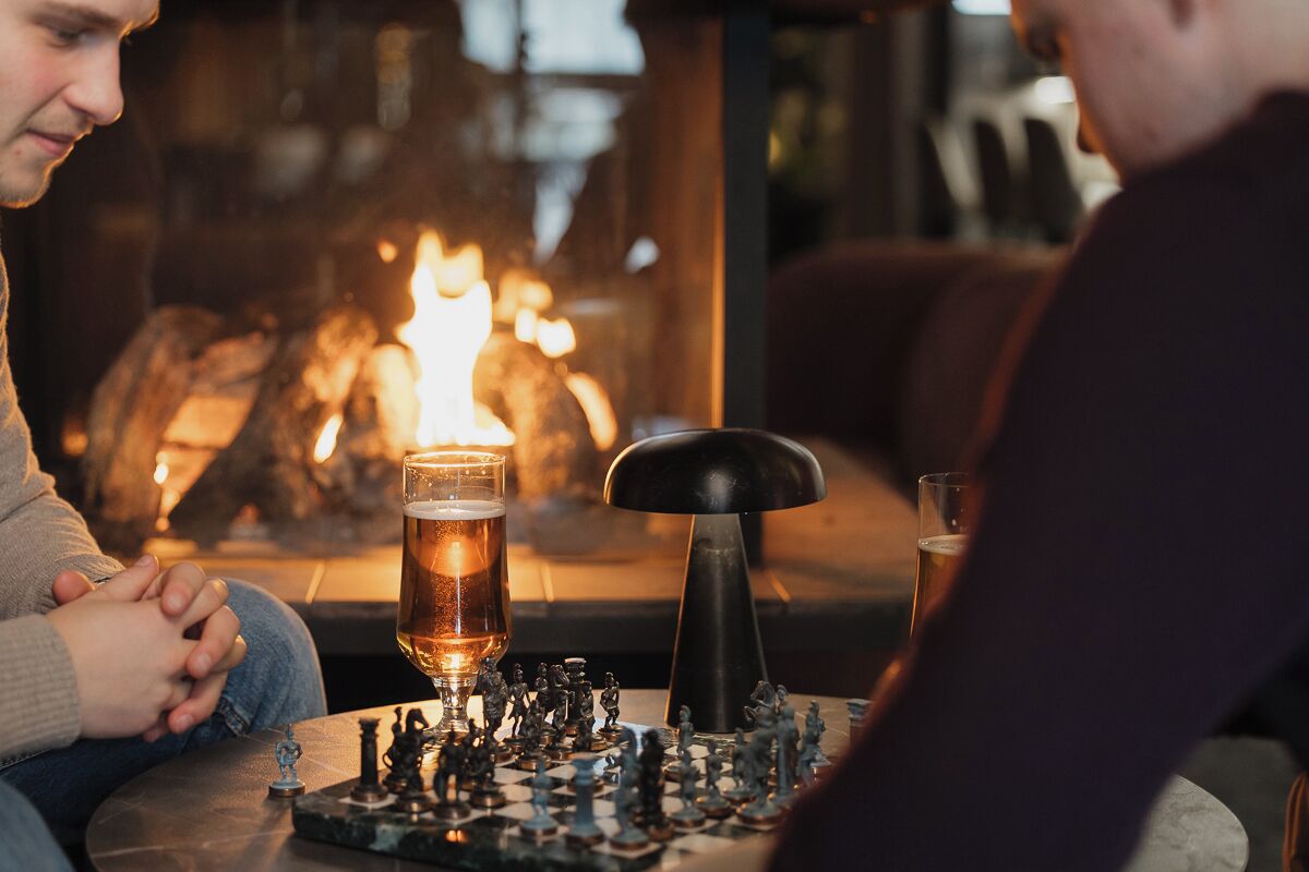 Ambiente acolhedor em interior com lareira, duas canecas de cerveja e um tabuleiro de xadrez.
