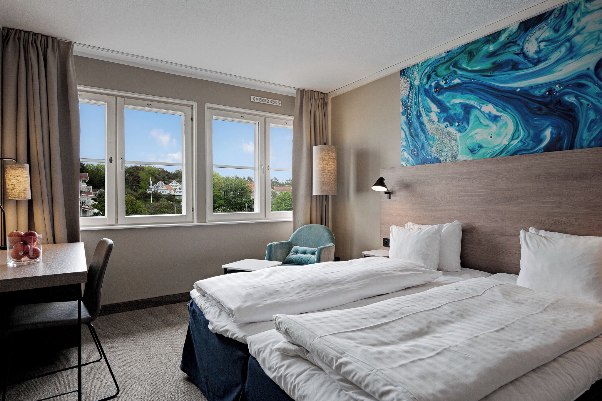 A hotel room with two beds, a desk, a chair, and a large window with a view of houses.