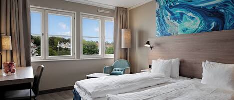 A hotel room with two beds, a desk, a chair, and a large window with a view of houses.