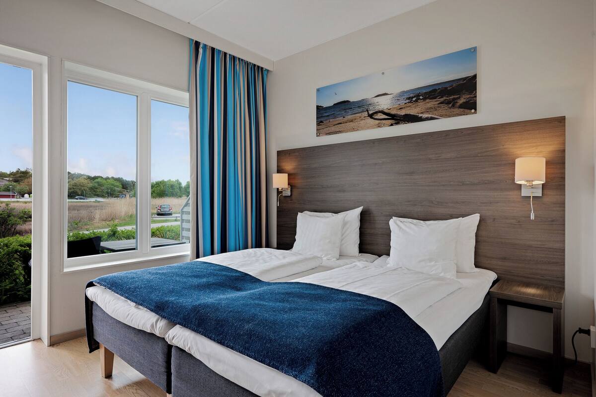 A hotel room with a large bed, a view of the outdoors, a painting on the wall, and a nightstand with a lamp.