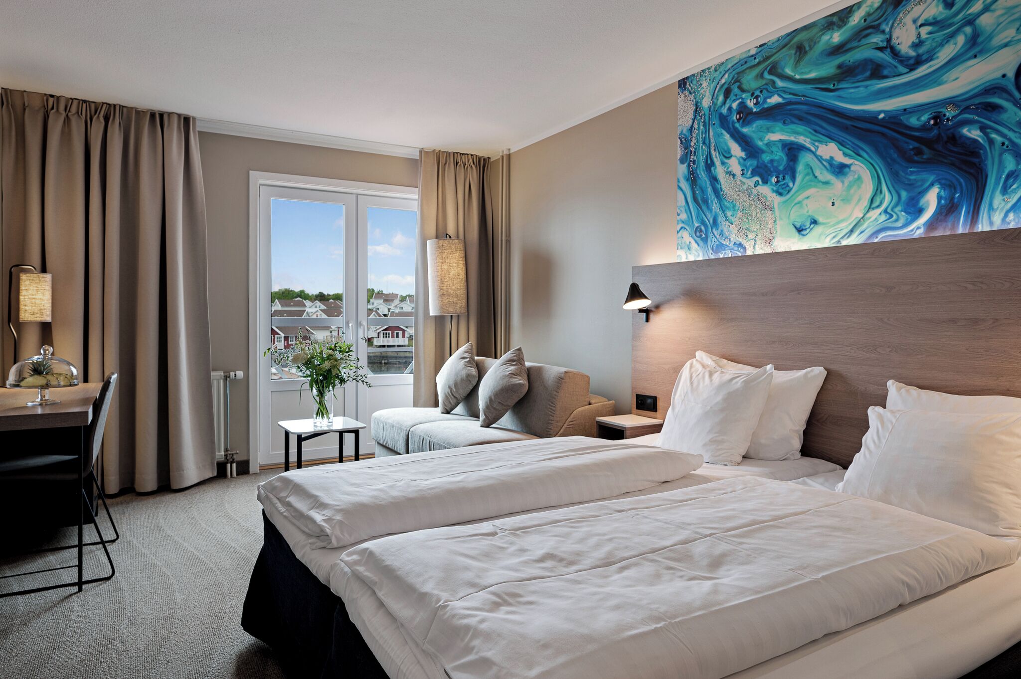 Habitación de hotel con dos camas, un escritorio, una silla, una lámpara, vistas al exterior y un cuadro abstracto grande en la pared.