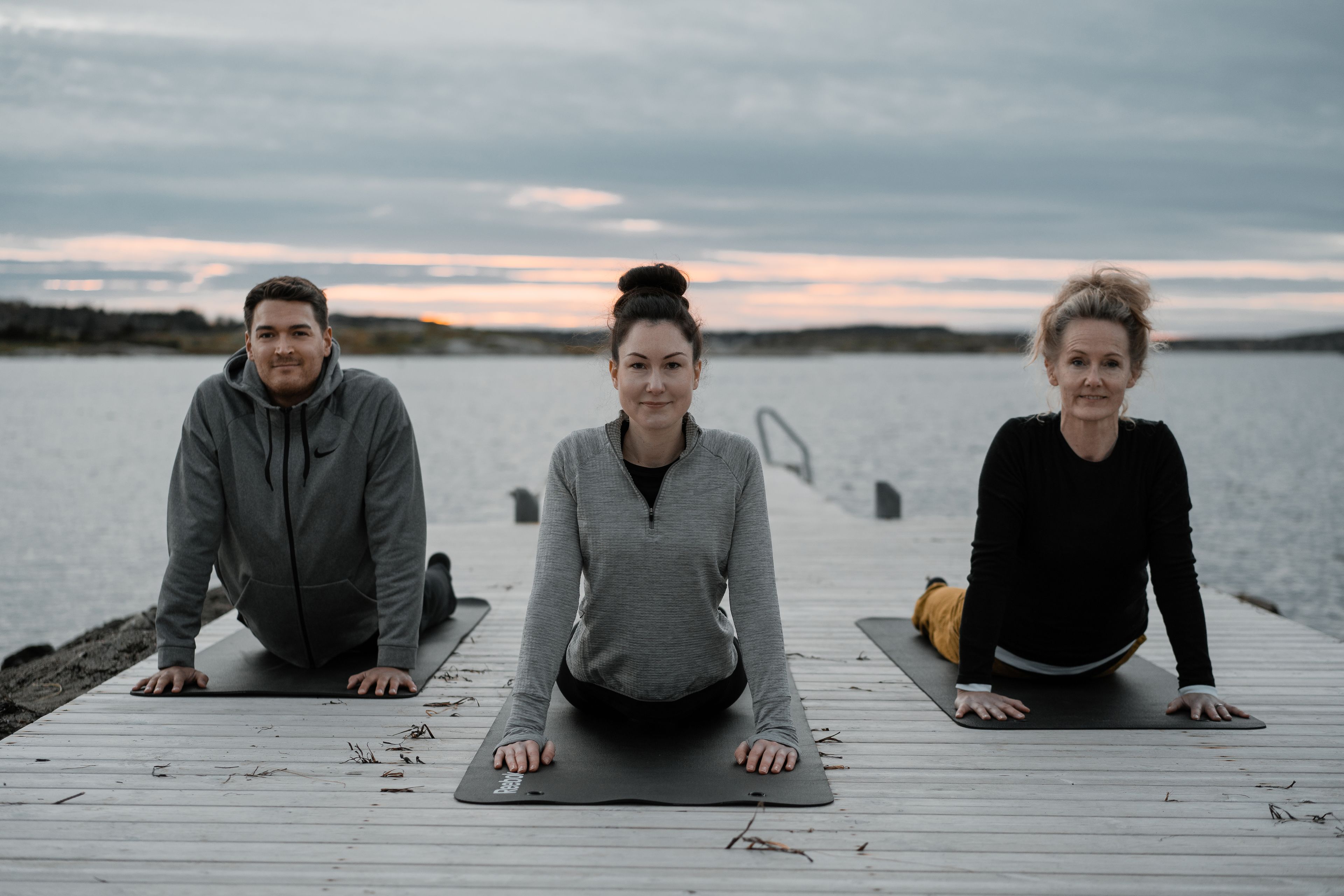Three people on yoga mats on a wooden dock by a body of water.