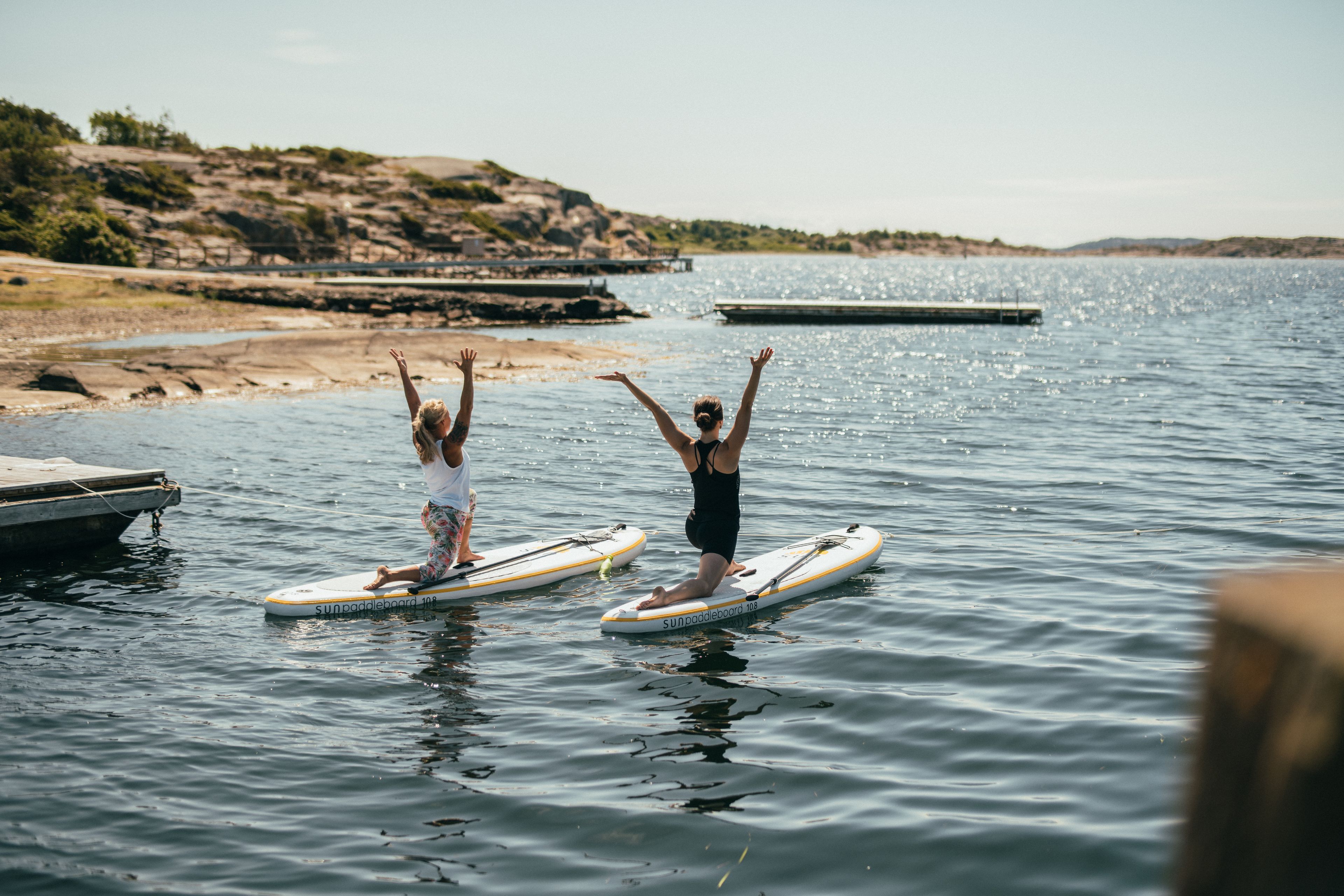 Two people on paddleboards in a calm body of water with a dock and rocky shoreline in the background.