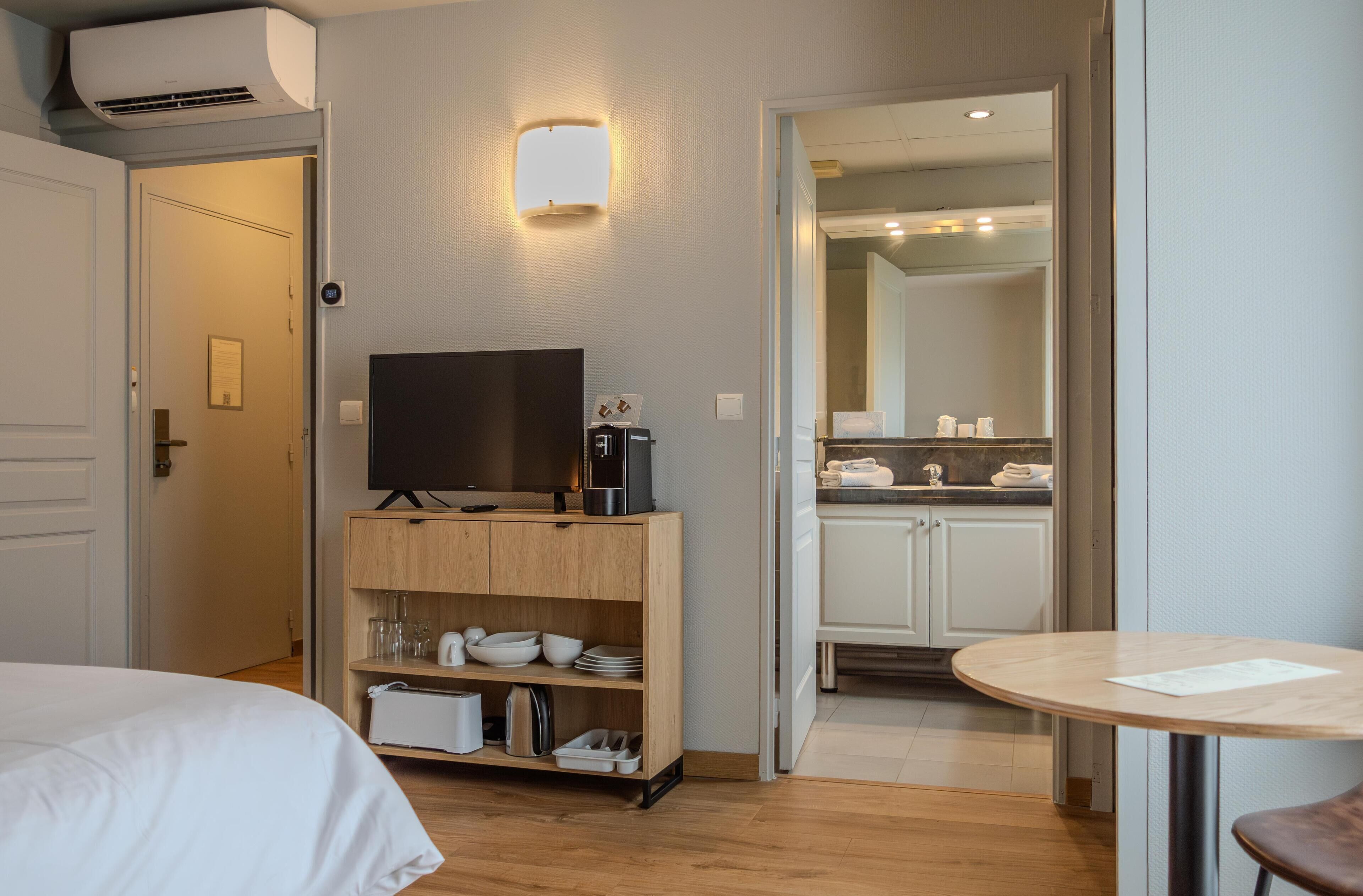 Et hotelværelse med en seng, et fjernsyn på et træstel, et lille køkkenområde med skabe og en vask, samt et rundt bord med en stol.