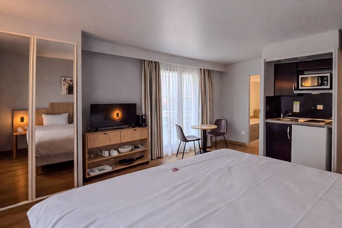 Quarto de hotel com cama, televisão, secretária com cadeira, área de cozinha e roupeiro com porta deslizante.