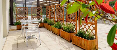 An outdoor patio with a table and chairs, wooden planters with plants, and a glass enclosure in the background.