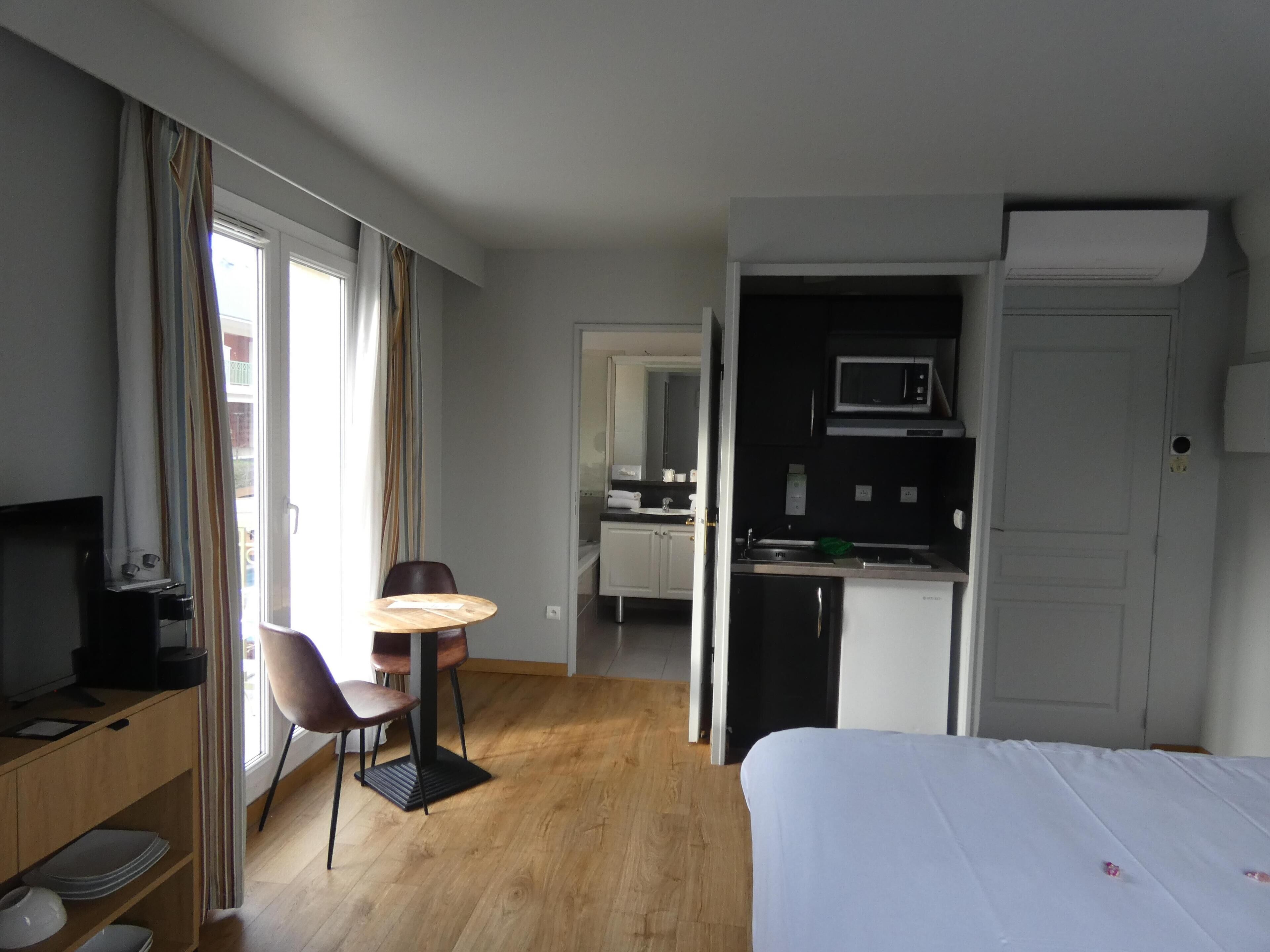A hotel room with a bed, a desk with a chair, a television, a dining table with chairs, a kitchen area with a microwave, and a sliding door leading to a bathroom.
