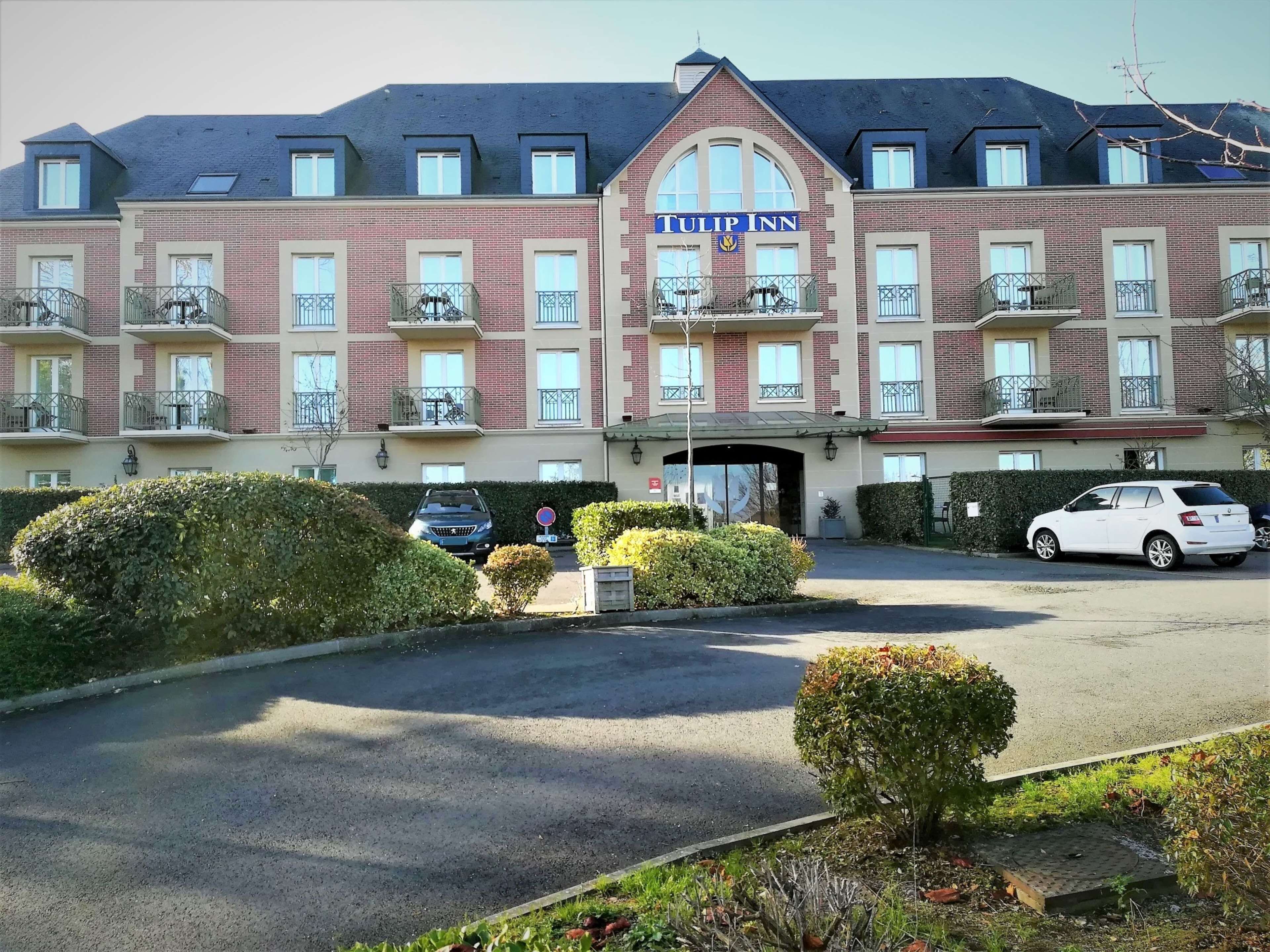 Un hôtel à plusieurs étages, reconnaissable à son grand panneau « Tulip Inn », doté de balcons à chaque étage, d’une entrée sécurisée par un portail et d’un espace de stationnement.