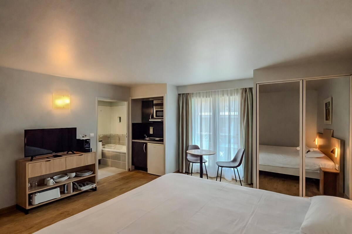 Habitación de hotel con cama, televisión, escritorio con silla, kitchenette pequeña y baño.