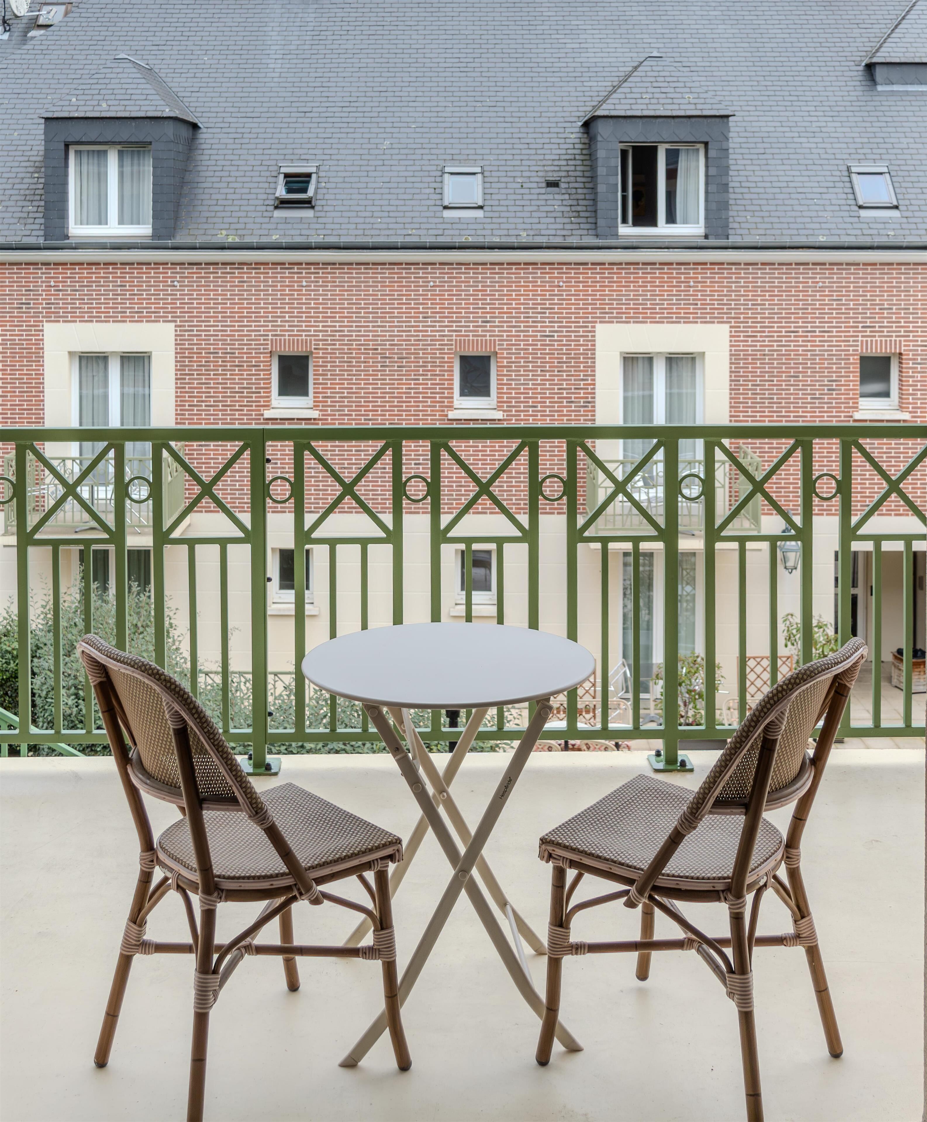 A balcony with two wicker chairs and a table, a green metal railing, and a red brick building in the background.