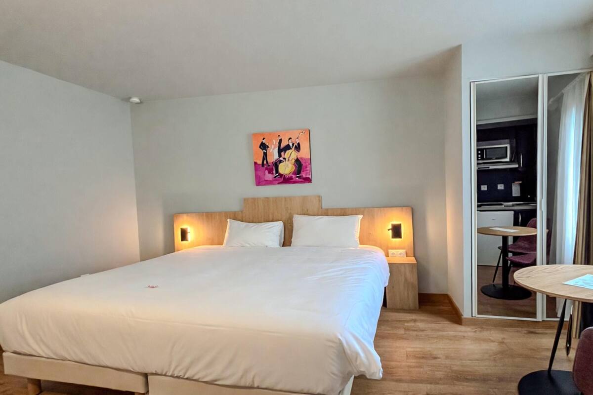 Quarto de hotel com cama grande, duas luminárias de cabeceira, um quadro na parede e uma cozinha americana visível através de uma porta aberta.