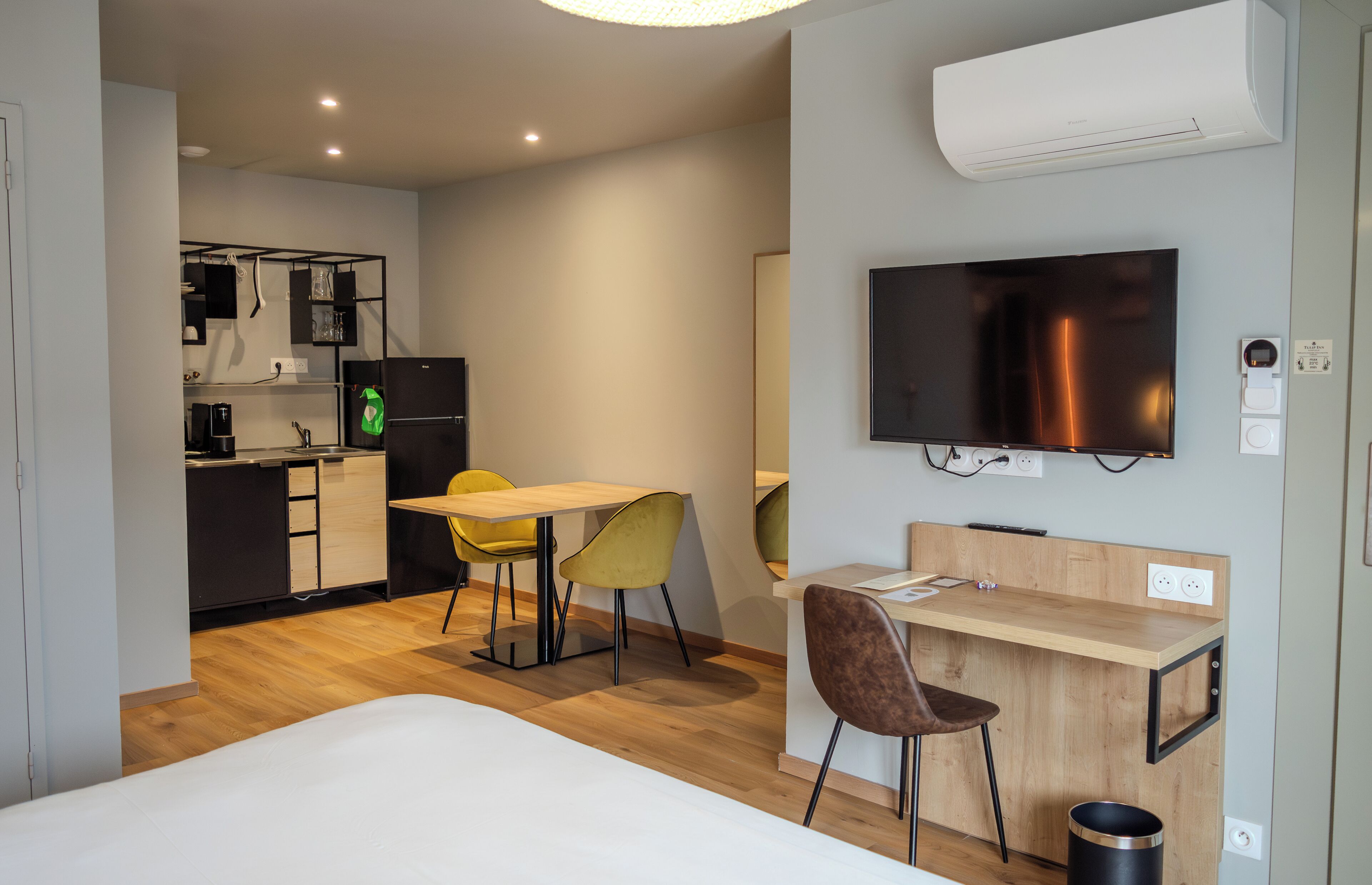 A modern hotel room with a bed, a desk with a chair, a television, and a kitchenette.