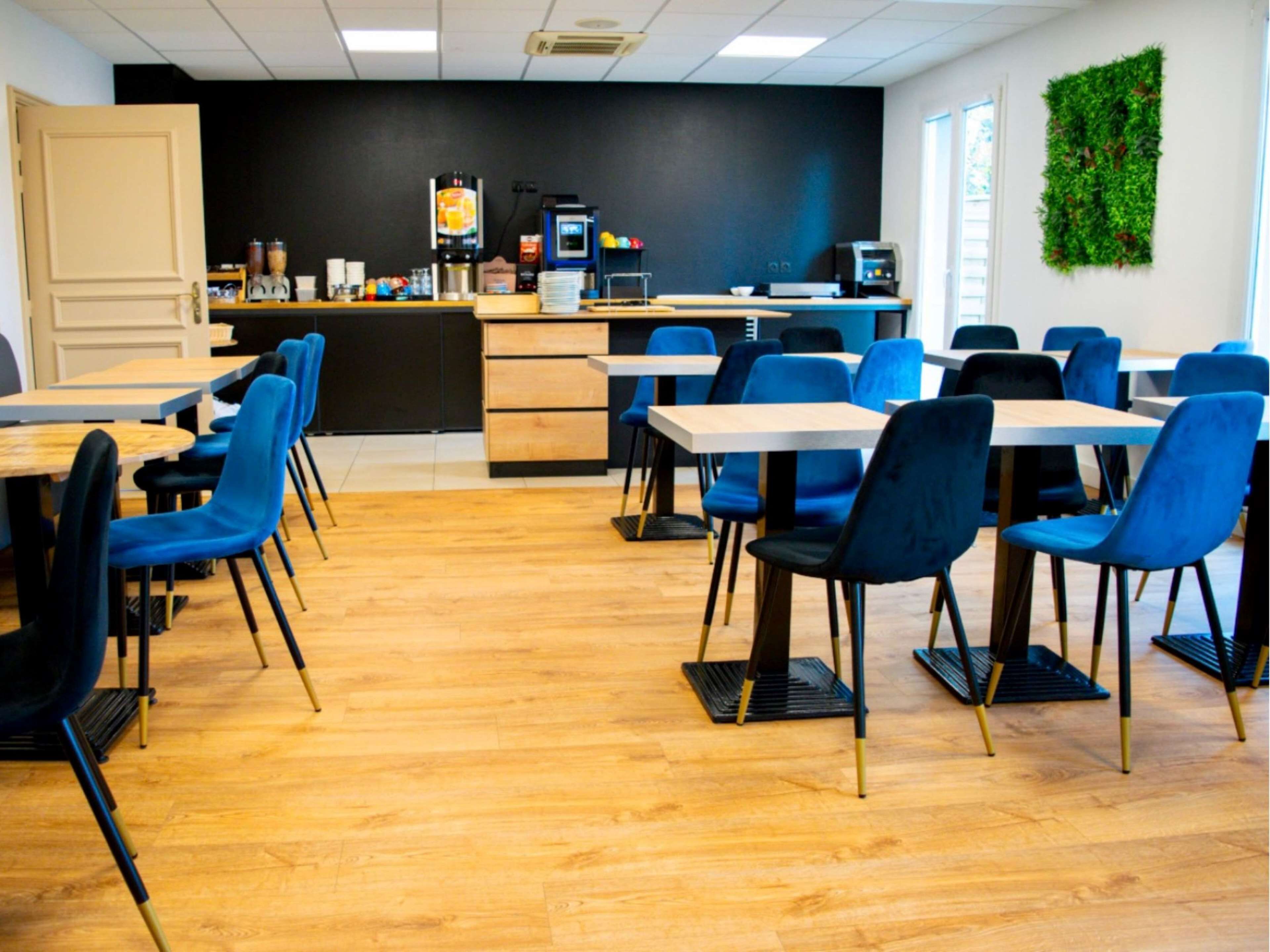 A modern dining area with wooden tables and blue chairs, a counter with a coffee machine, and a green wall decoration.