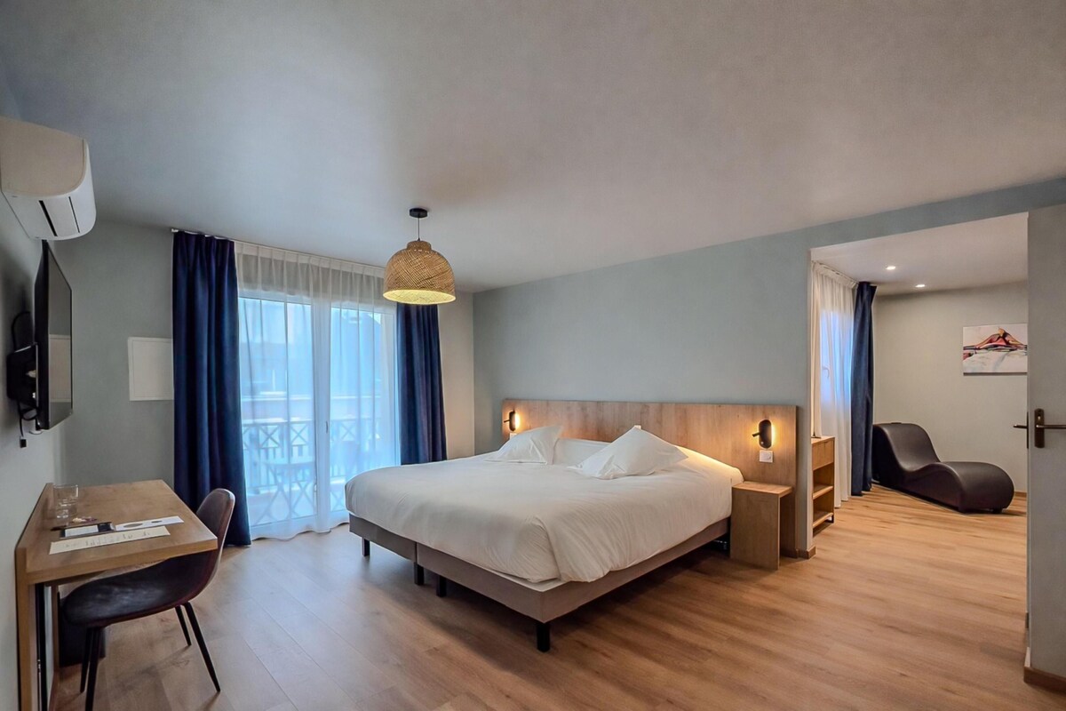 Habitación de hotel con cama, mesitas de noche, escritorio con silla, televisión y balcón con cortinas.