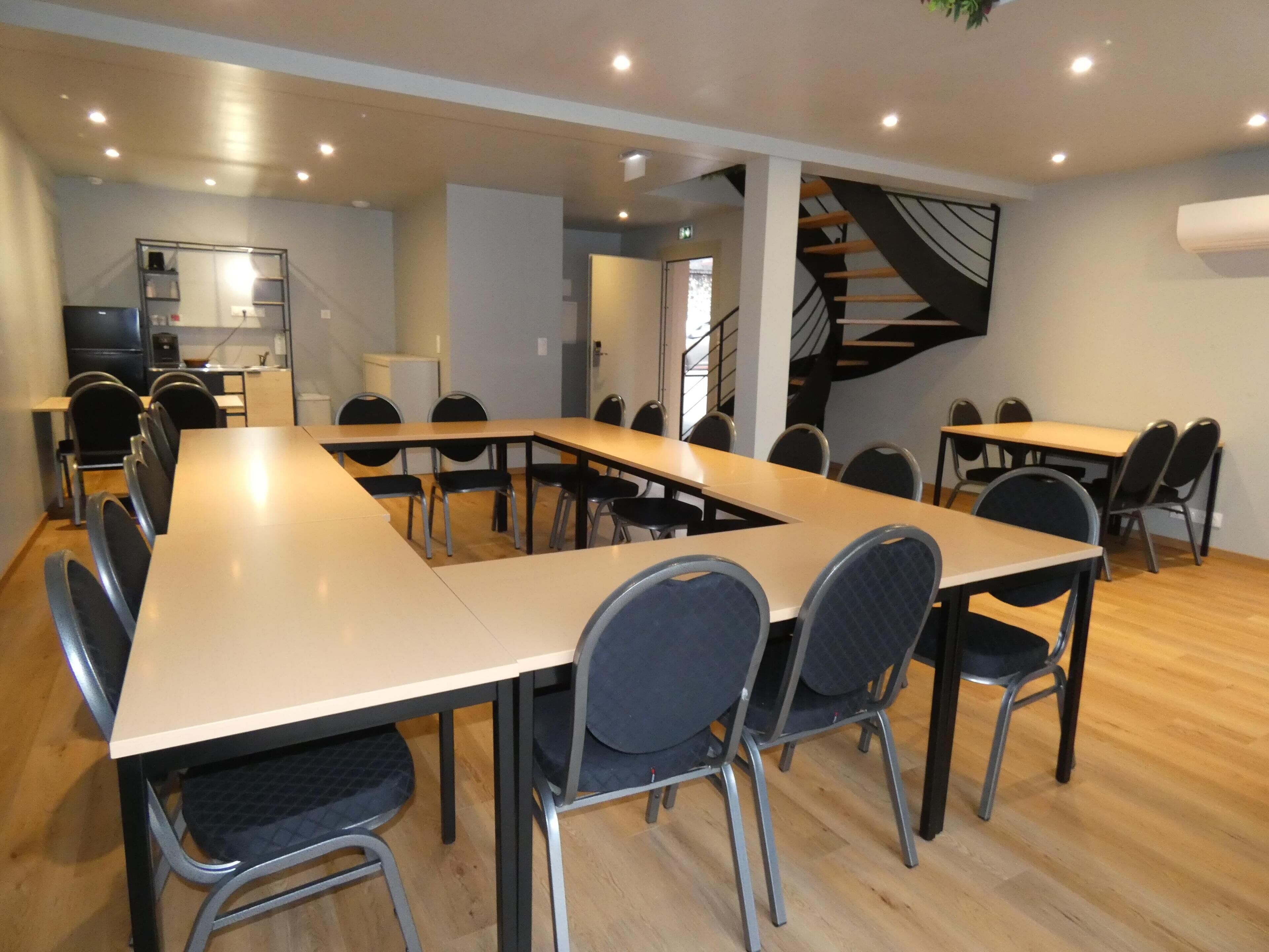 A modern meeting room with a long table and chairs, a kitchen area, and a staircase.