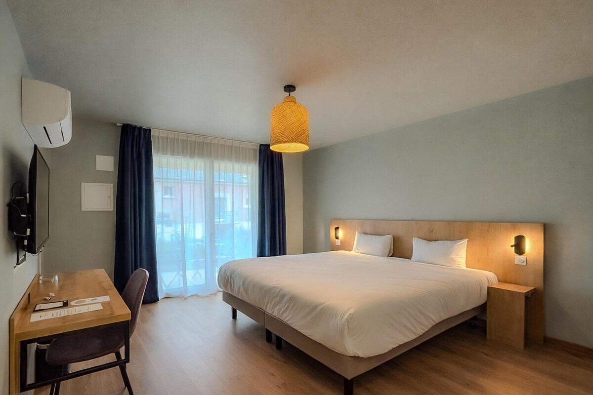 Habitación de hotel con una cama grande, un escritorio con silla, televisor y ventana con cortinas.