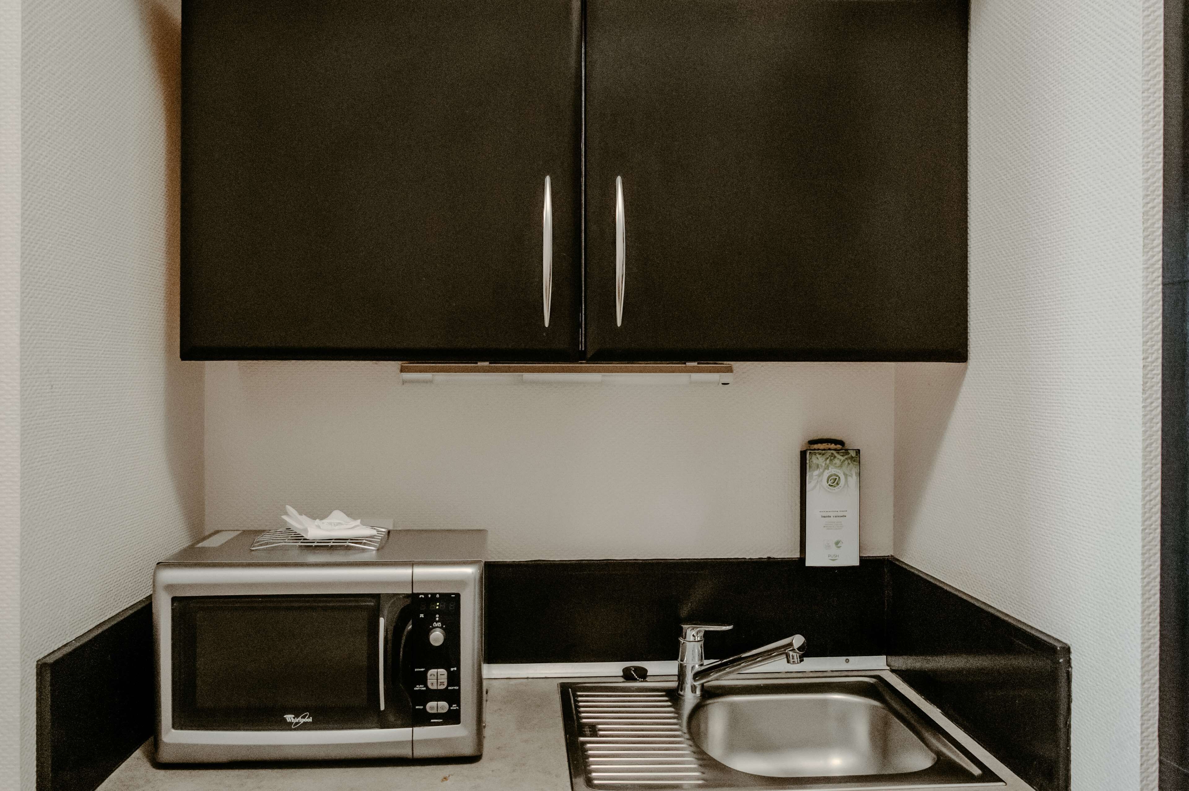 Une cuisine de taille réduite, équipée d’un four à micro-ondes, d’un évier et de meubles de rangement de couleur sombre.