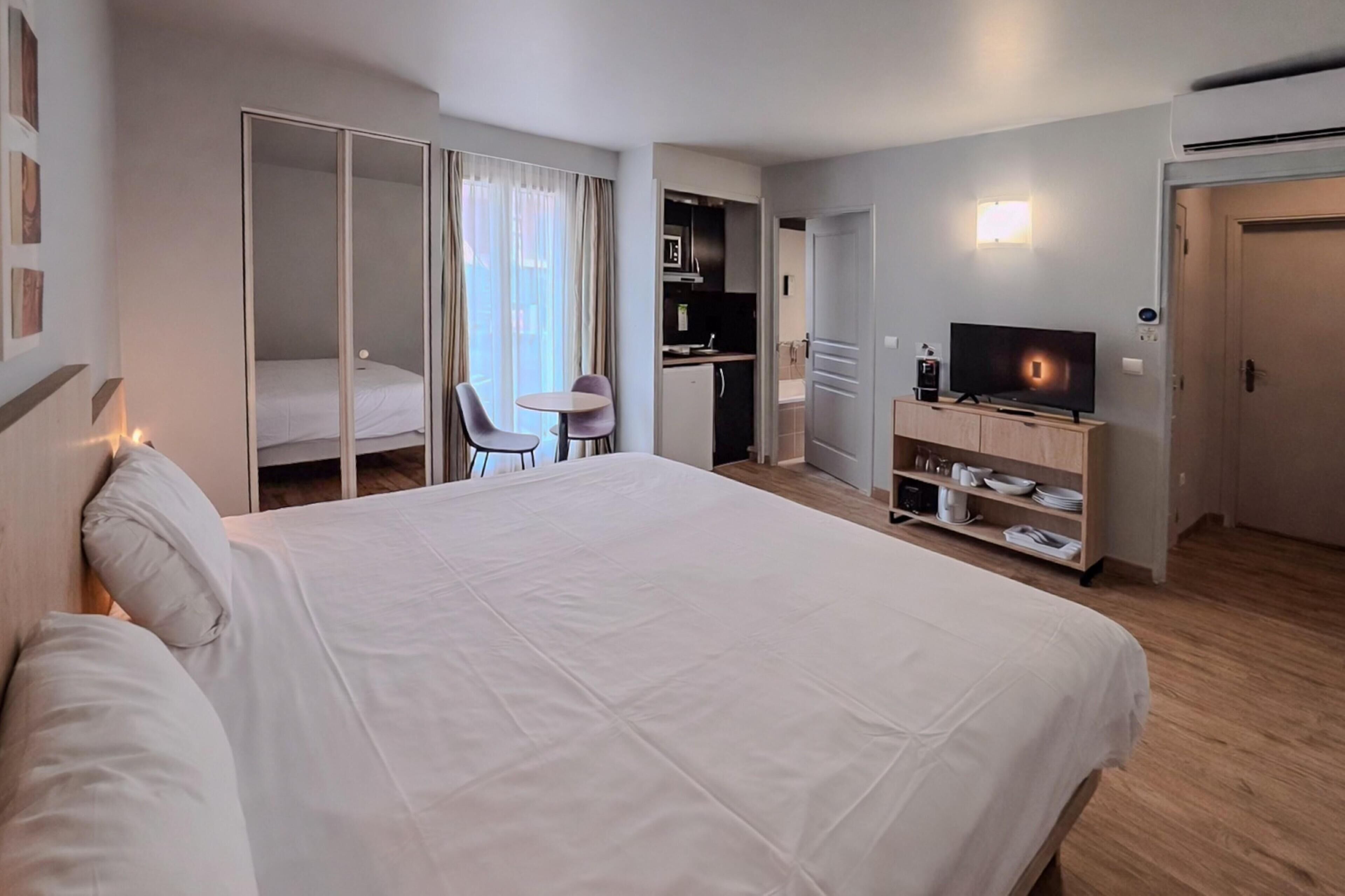 A hotel room with a large bed, a desk with a chair, a kitchenette, and a TV.