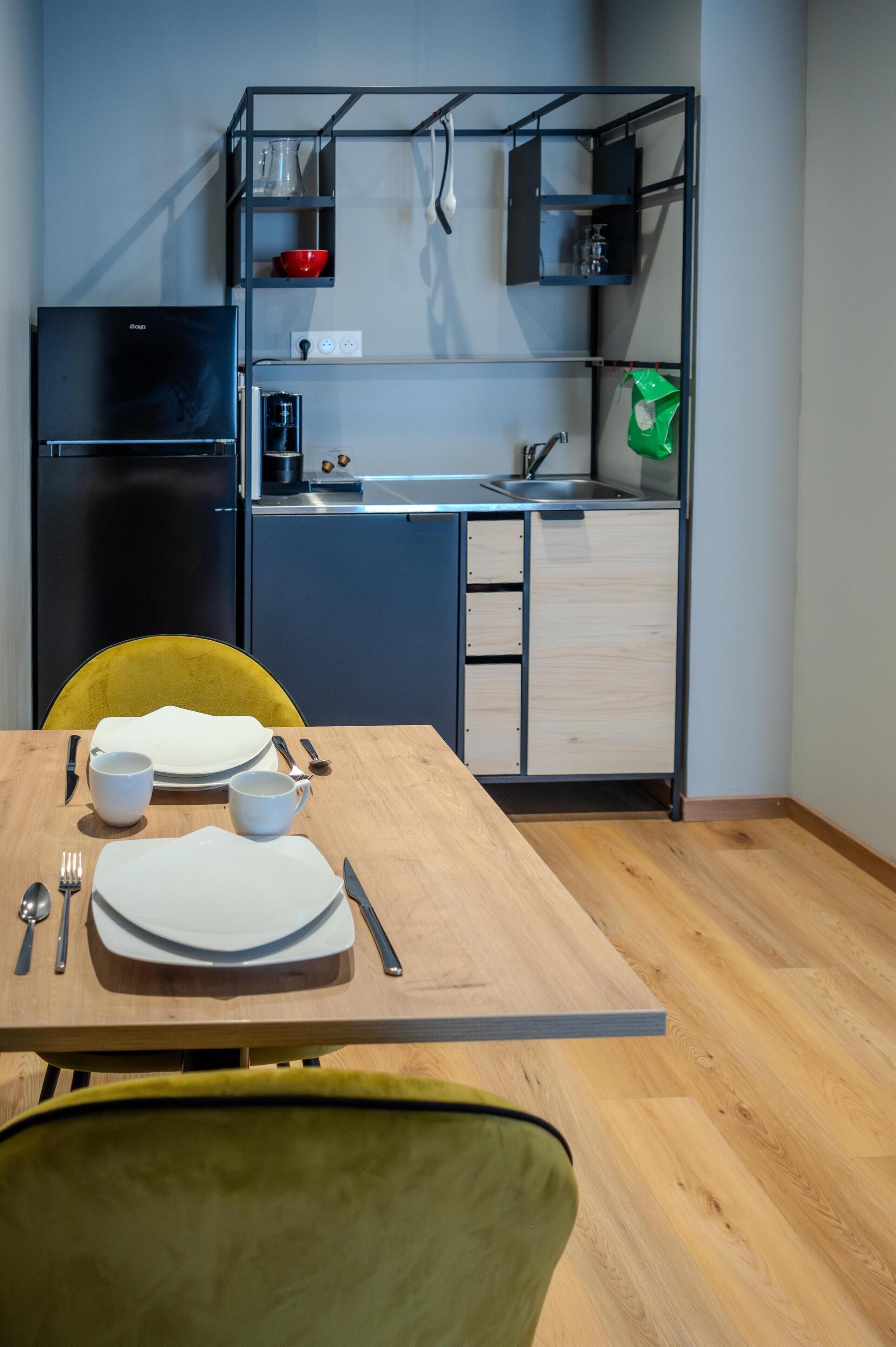 Une cuisine compacte avec un réfrigérateur noir, une table à manger en bois et des chaises jaunes, ainsi qu’une étagère intégrée au-dessus du plan de travail.