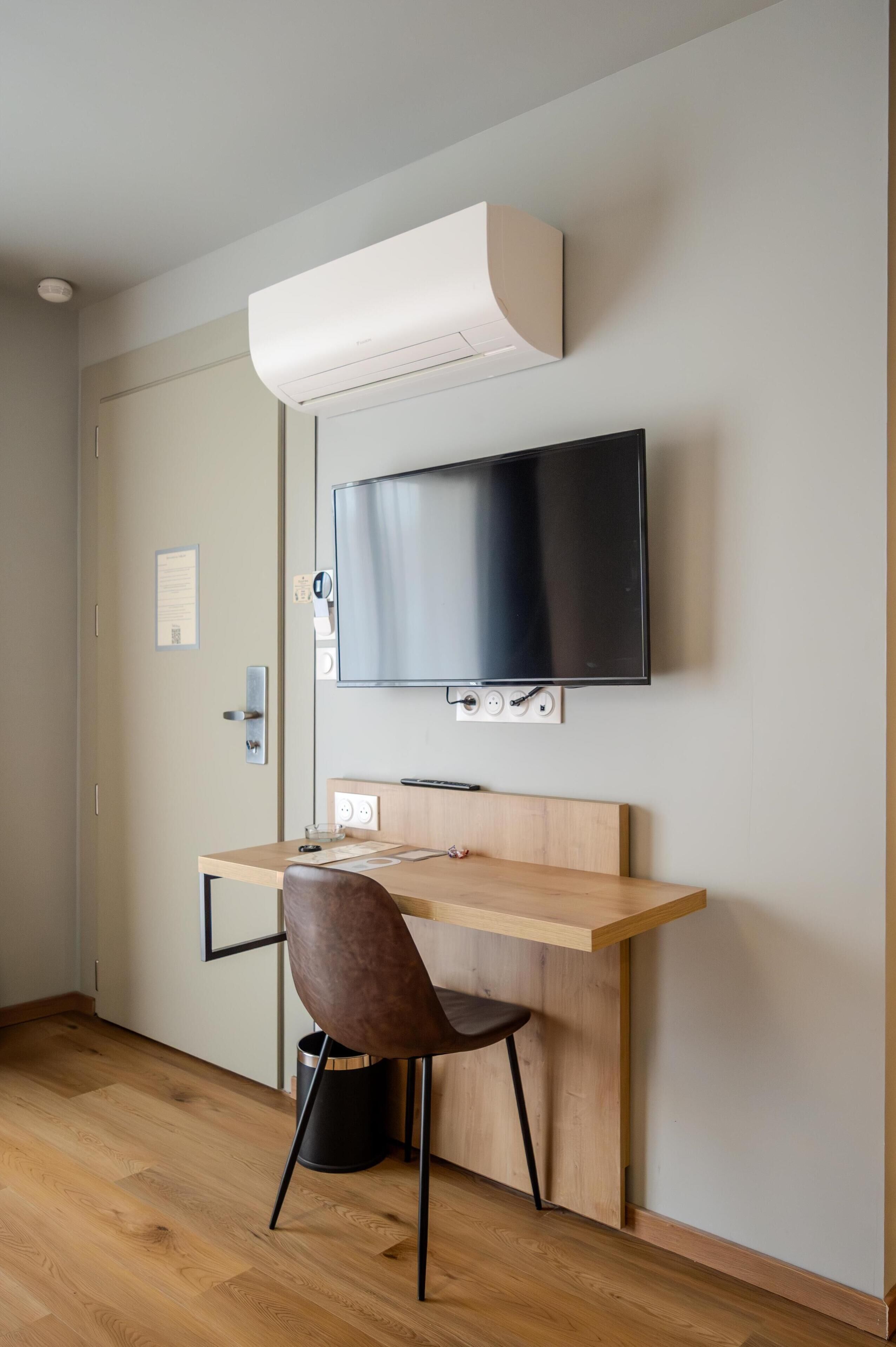 Una habitación moderna con un escritorio de madera, una silla marrón, un televisor de pantalla plana montado en la pared y aire acondicionado en el techo.