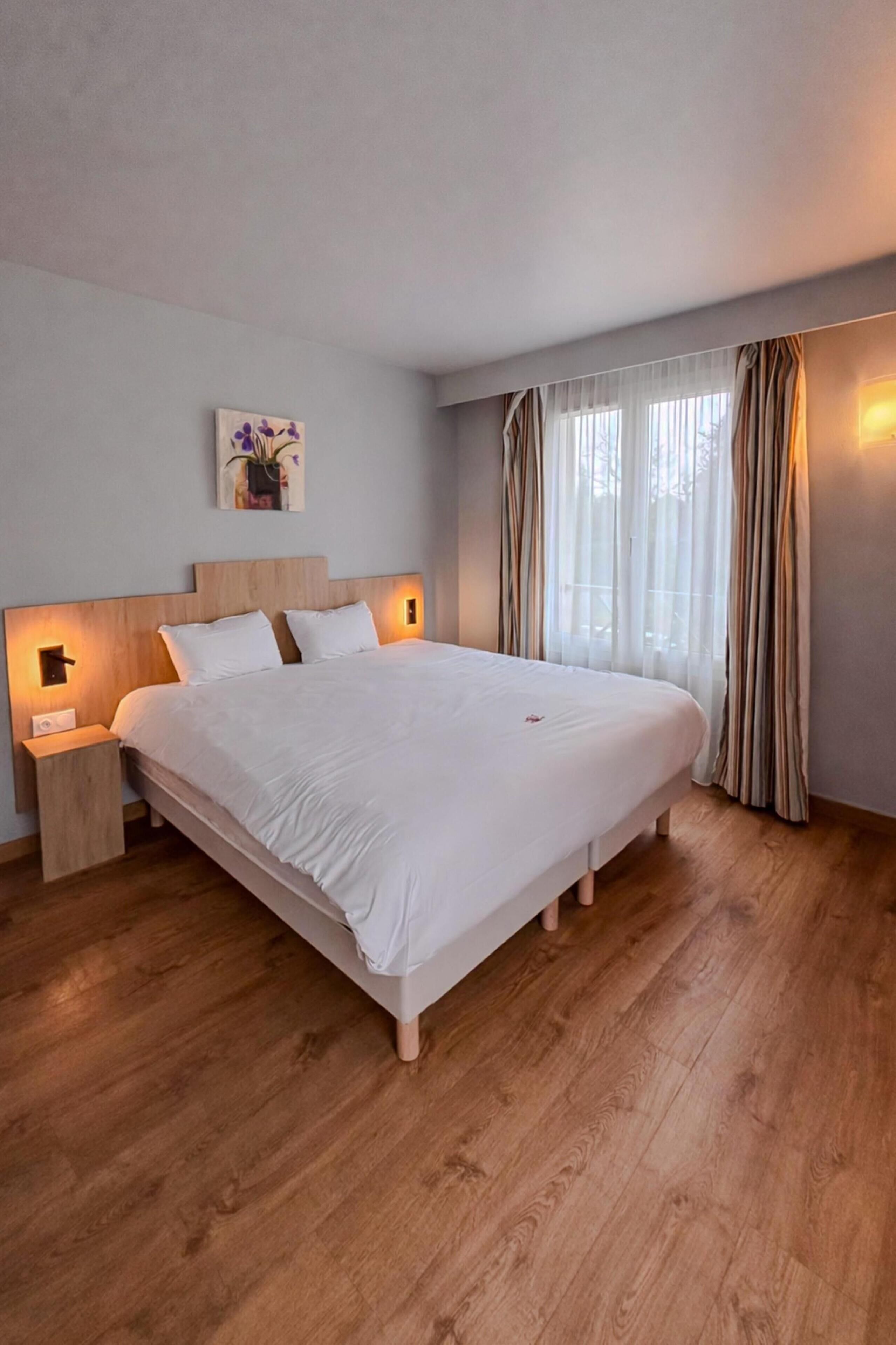 A large bed with a wooden headboard, two bedside tables with lamps, a painting on the wall, and a window with curtains.