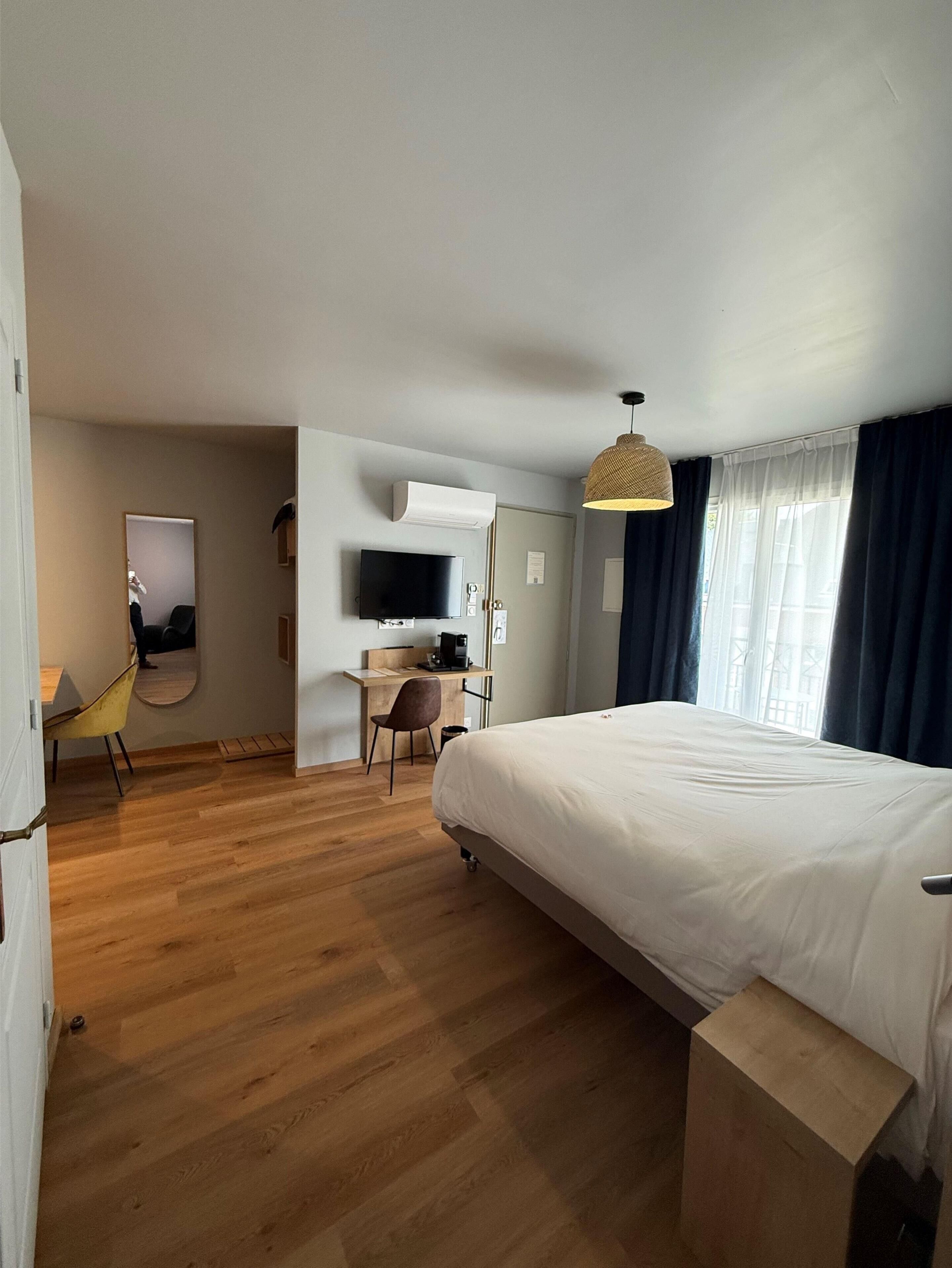 A modern hotel room with a large bed, a desk with a chair, a television, and a window with curtains.