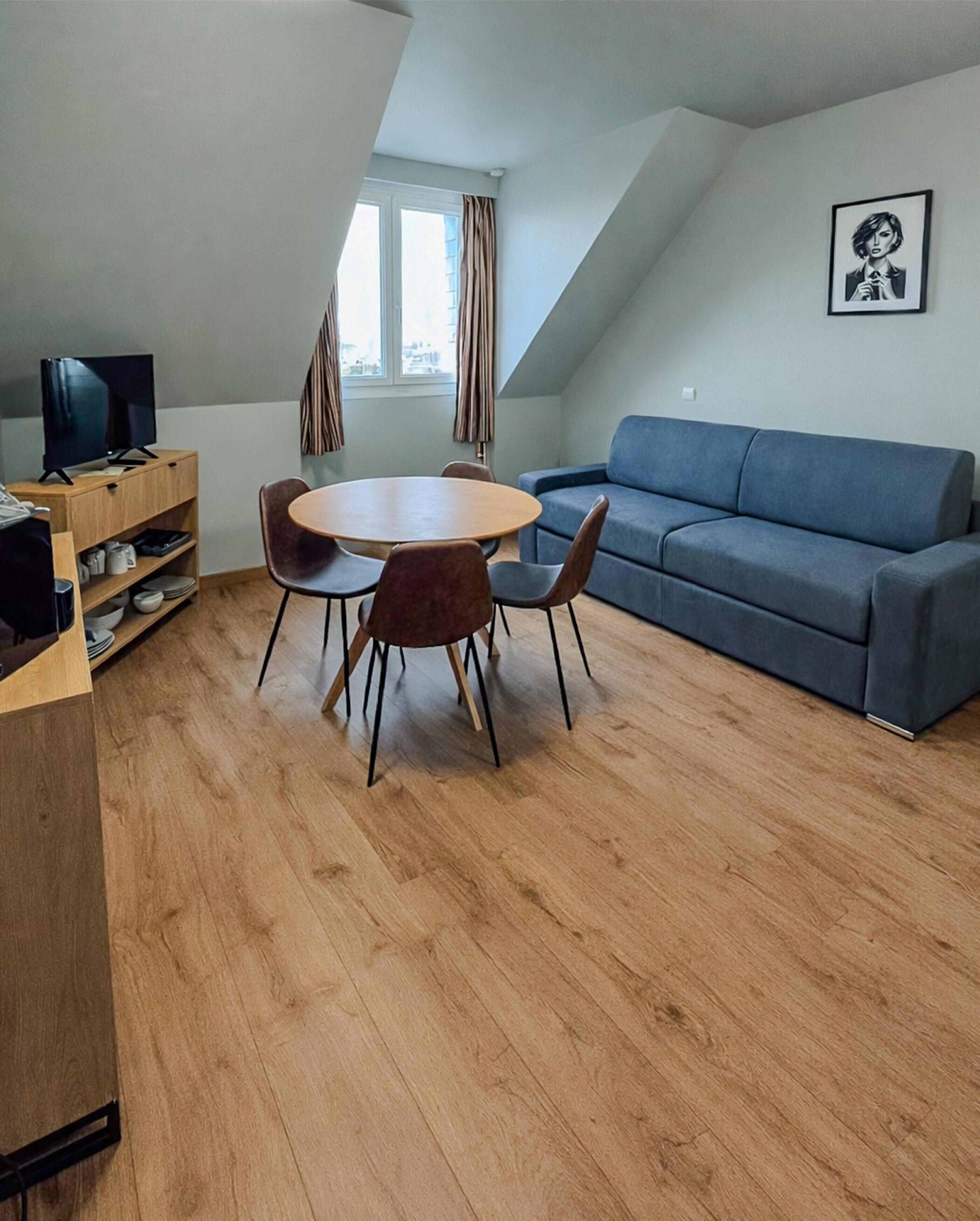 A modern living room with a round wooden table, four chairs, a blue sofa, and a television on a wooden stand.