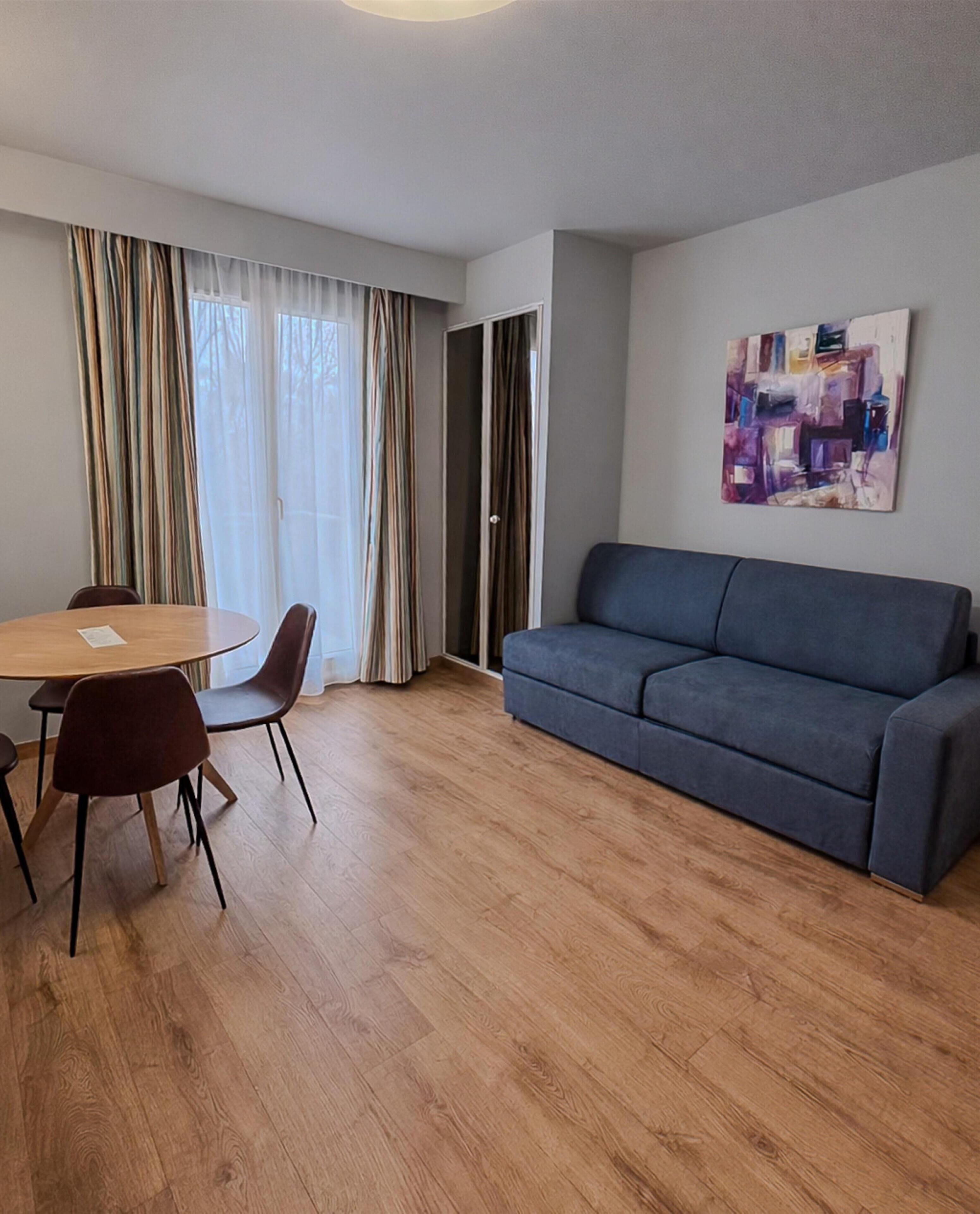 Una sala de estar moderna con un sofá gris, una mesa de comedor de madera con sillas y un ventanal con cortinas.