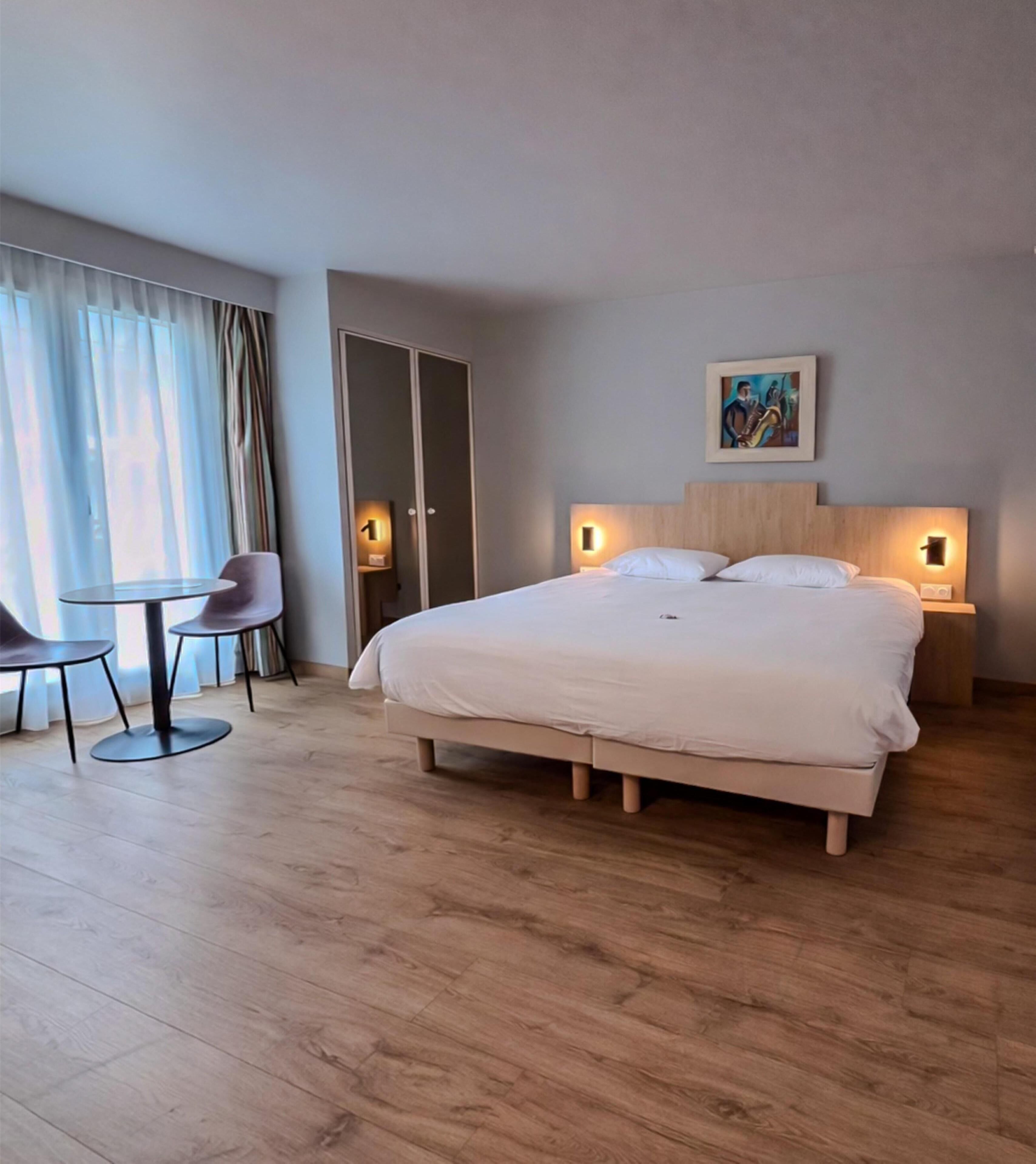 Una habitación de hotel moderna con una cama grande, una pequeña mesa redonda con sillas, un armario y un cuadro en la pared.