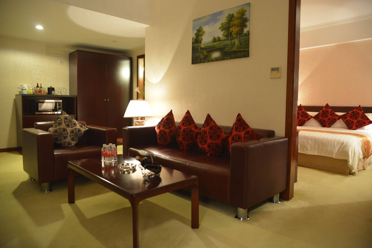 A hotel room with a brown leather sofa, a wooden coffee table, a television, and a bed with red patterned pillows.