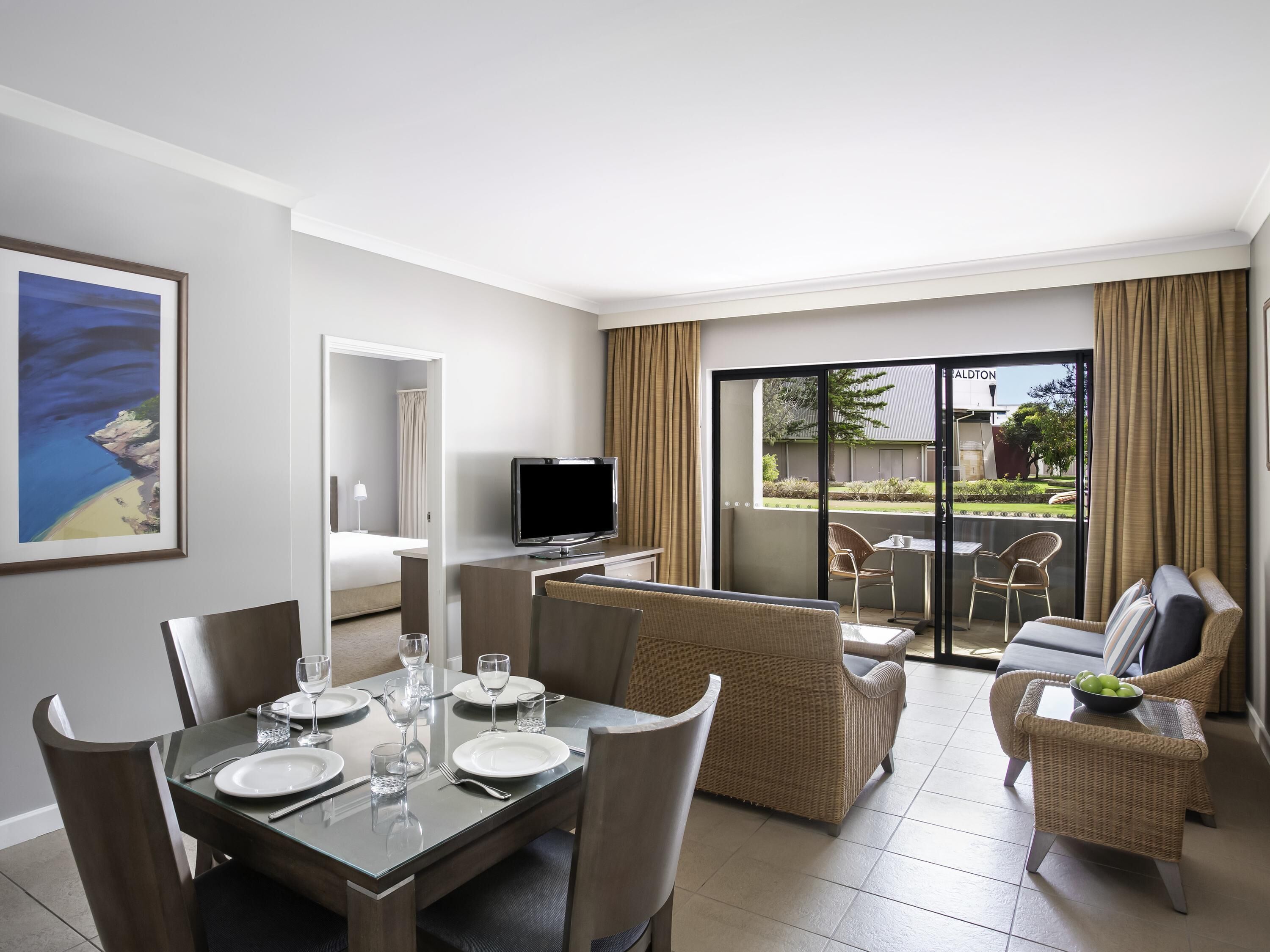 A hotel room with a dining area, a television, a balcony with a view of the outside, and a bed.
